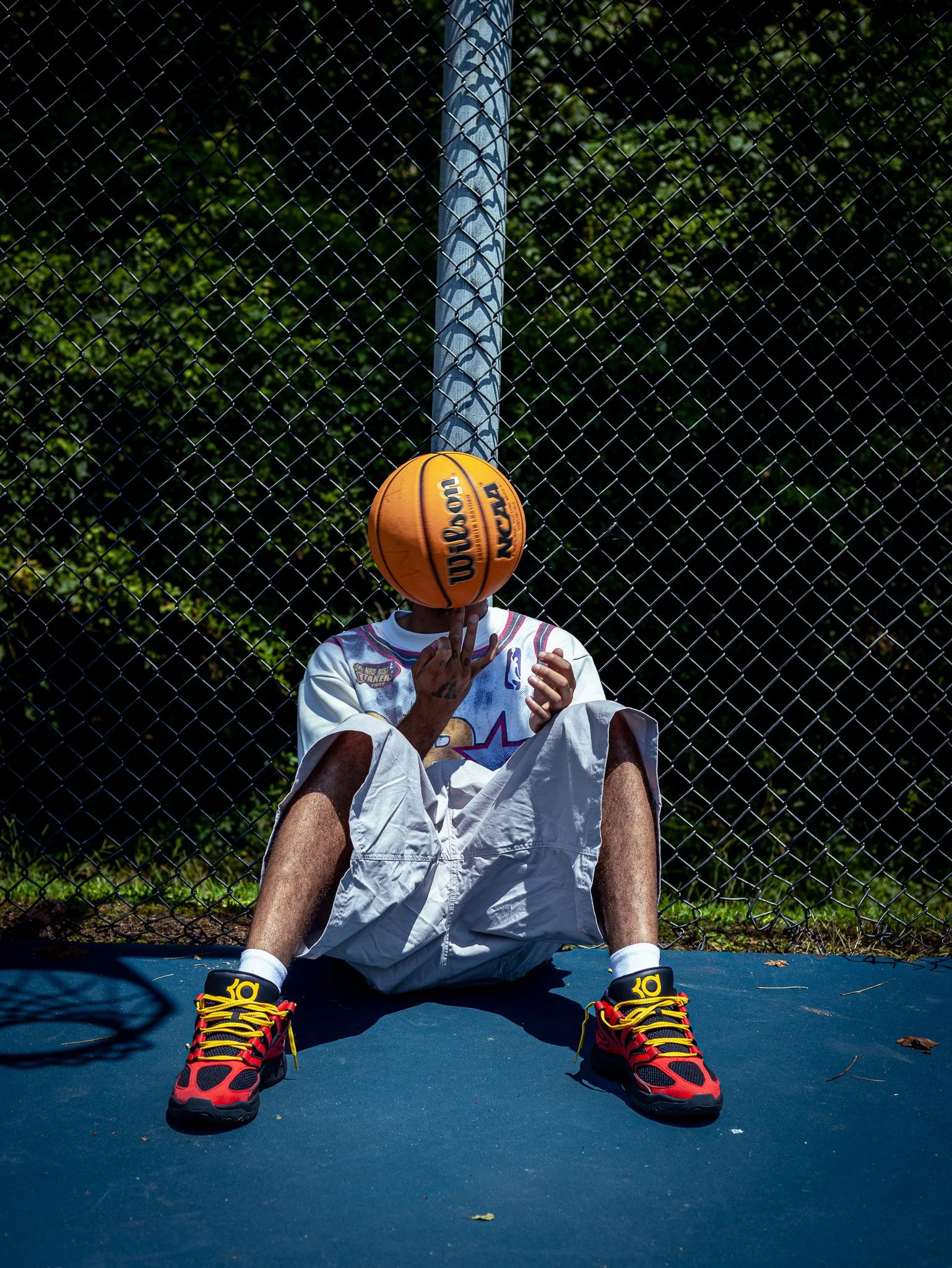 Person sitting on a basketball court with a basketball head, wearing sneakers, shorts, and a jersey, behind a chain-link fence, with trees in the background.
