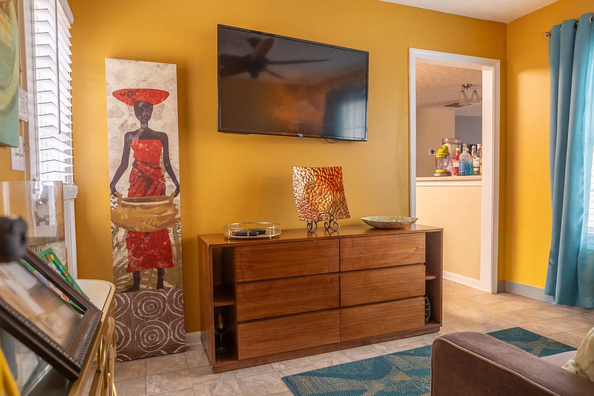 Living room with yellow walls, a flat-screen TV on the wall, a wooden TV stand with decorative items, a colorful lamp, and a tall artwork of a woman carrying a basket. There is a doorway to the kitchen with bottled drinks and a blender, a window with