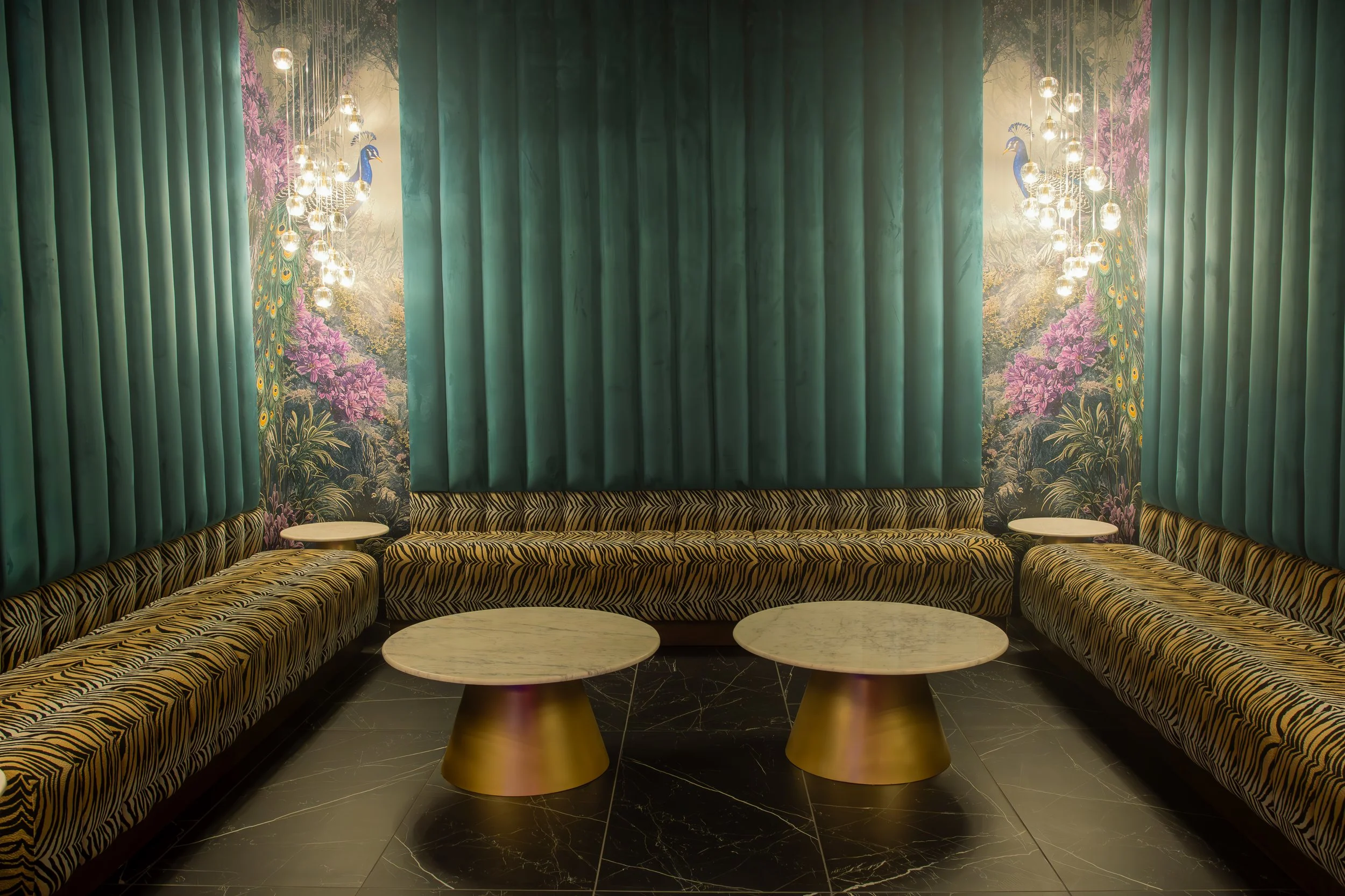 A luxurious lounge with zebra-print sofas arranged in a U-shape, gold and marble accent tables in the center, dark polished tile flooring, teal velvet curtains, a scenic peacock mural with purple flowers, and modern hanging light fixtures with expose