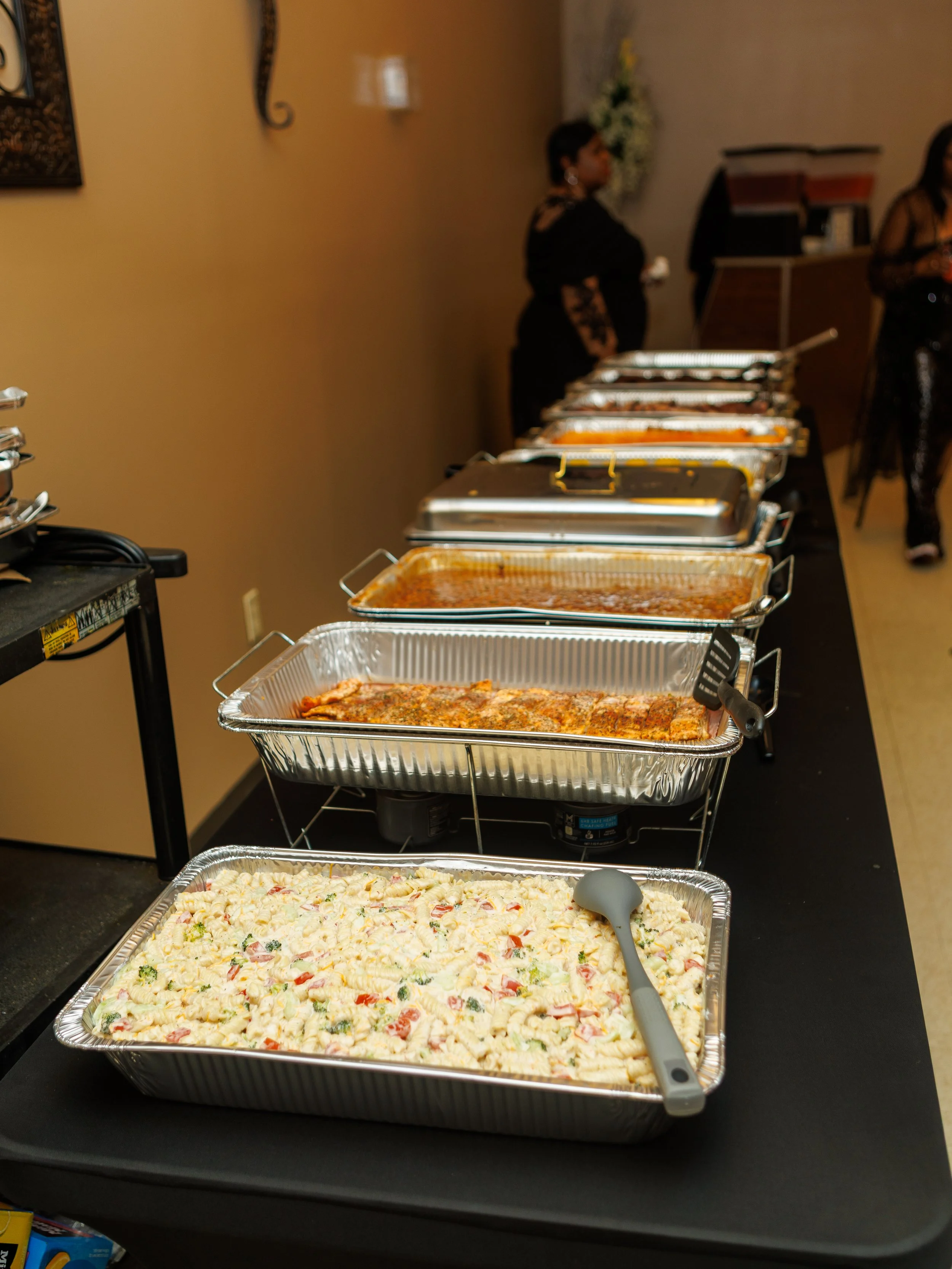 Trays of various foods arranged on a buffet table, including a salad, pasta, grilled meat, and other dishes, with people in the background preparing or serving food.