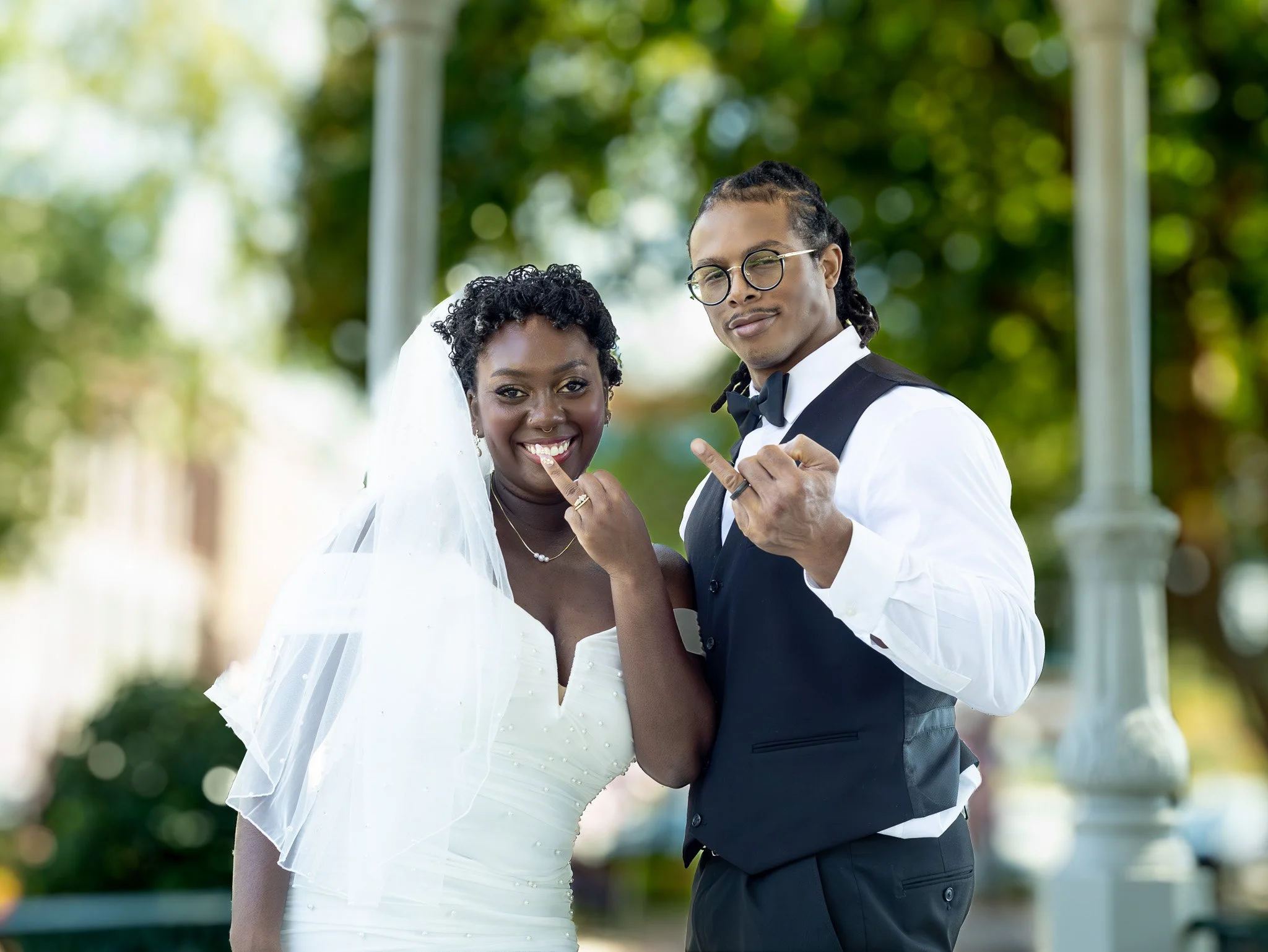 A newlywed couple in wedding attire making hand gestures, standing outdoors with trees in the background.