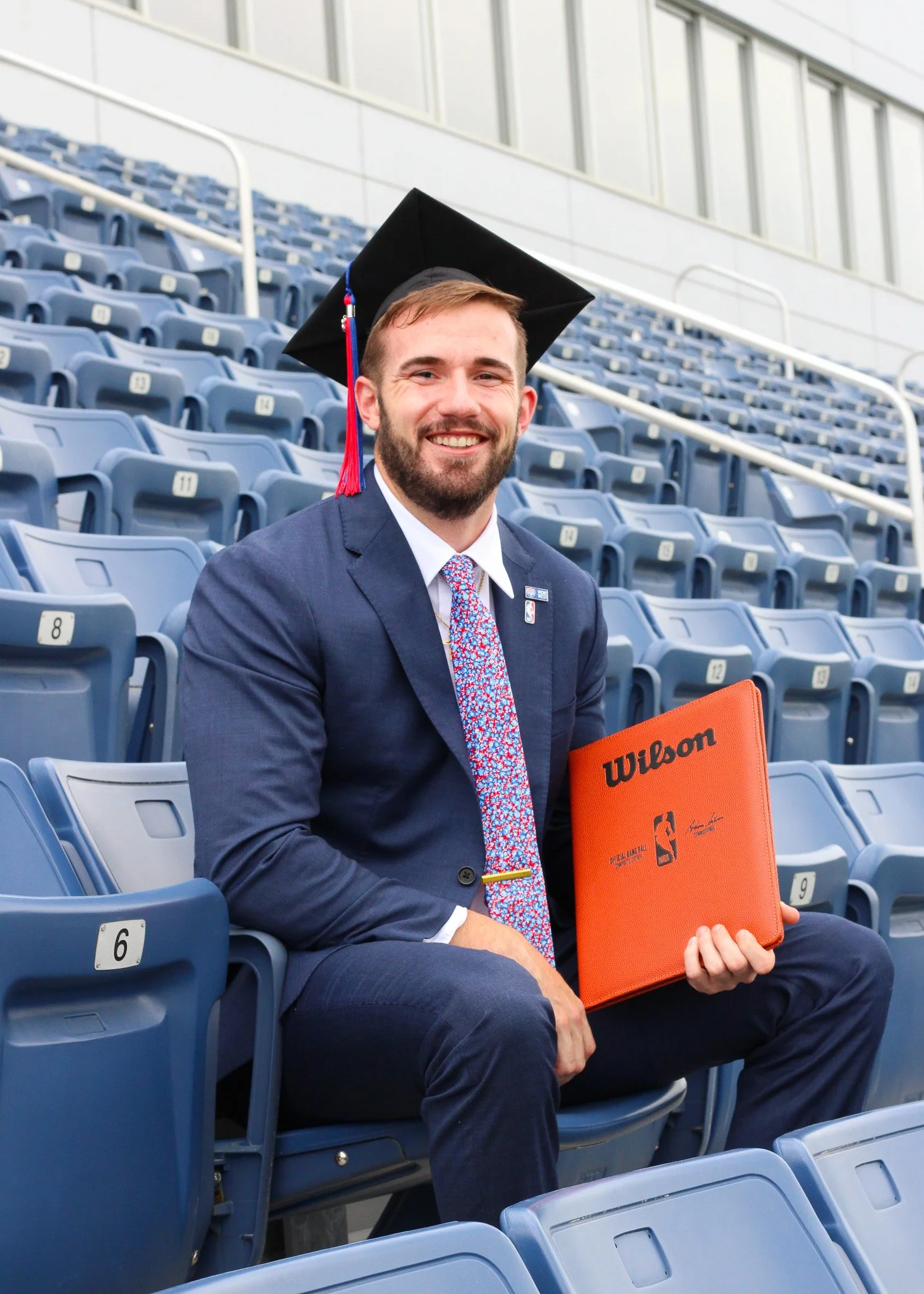 A man in a suit and tie, wearing a graduation cap, sitting in stadium seating with empty blue chairs, holding an orange Wilson binder.