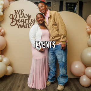A smiling couple posing at an event with a backdrop that says, 'We Can Bearly Wait,' surrounded by beige, blush, and cream balloons. The woman is wearing a pink dress and white blazer, and the man is wearing a tan jacket and jeans.