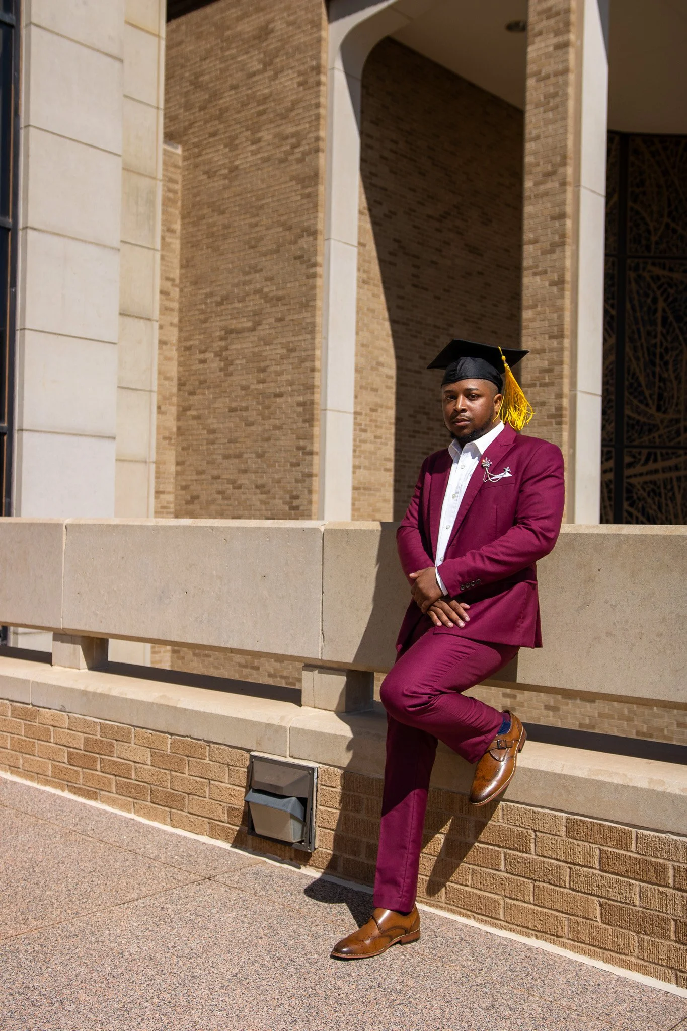 A young man in a maroon suit and white shirt wearing a graduation cap with a yellow tassel, standing outside a modern brick building, leaning against a beige stone ledge.