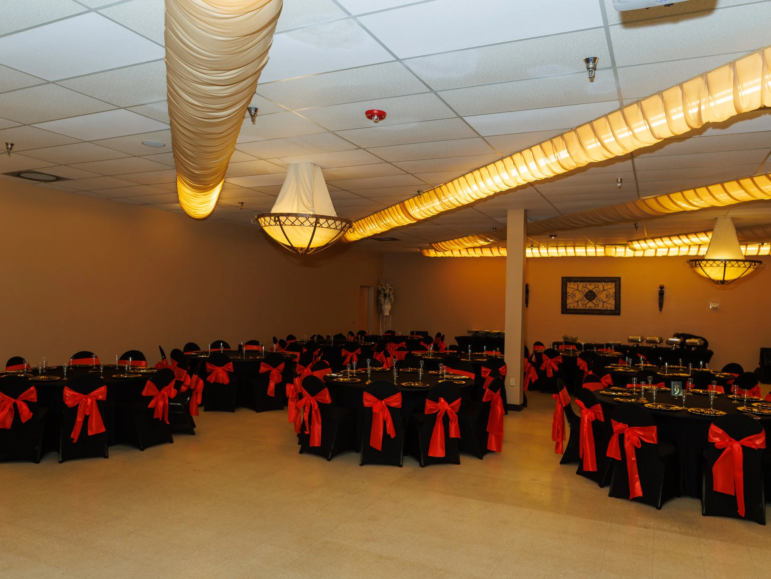 Ballroom decorated for a party with black tables and chairs adorned with red ribbons, set with dinnerware, under warm lighting and draped ceiling decorations.