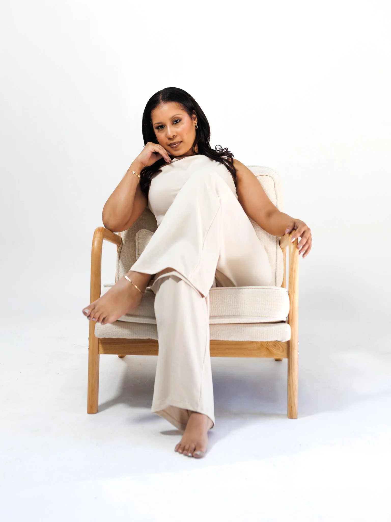 A woman sitting on a wooden chair with a beige cushion, wearing cream-colored pants and a matching top, with long dark hair, jewelry, and a confident expression, against a plain white background.