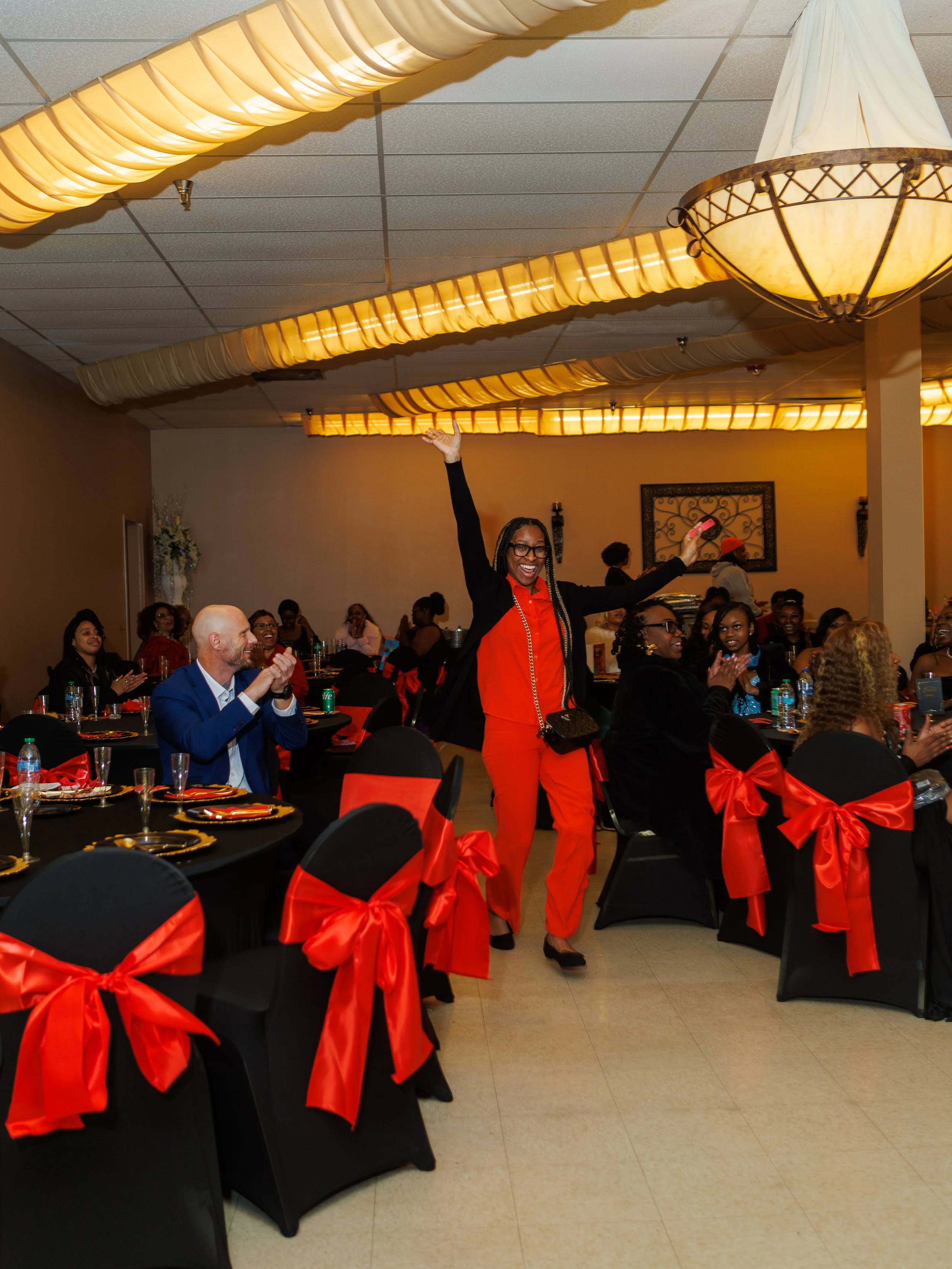 A woman in a red outfit with black blazer and glasses raising her arms joyfully in a banquet hall decorated with black chairs tied with red ribbons, and guests clapping.