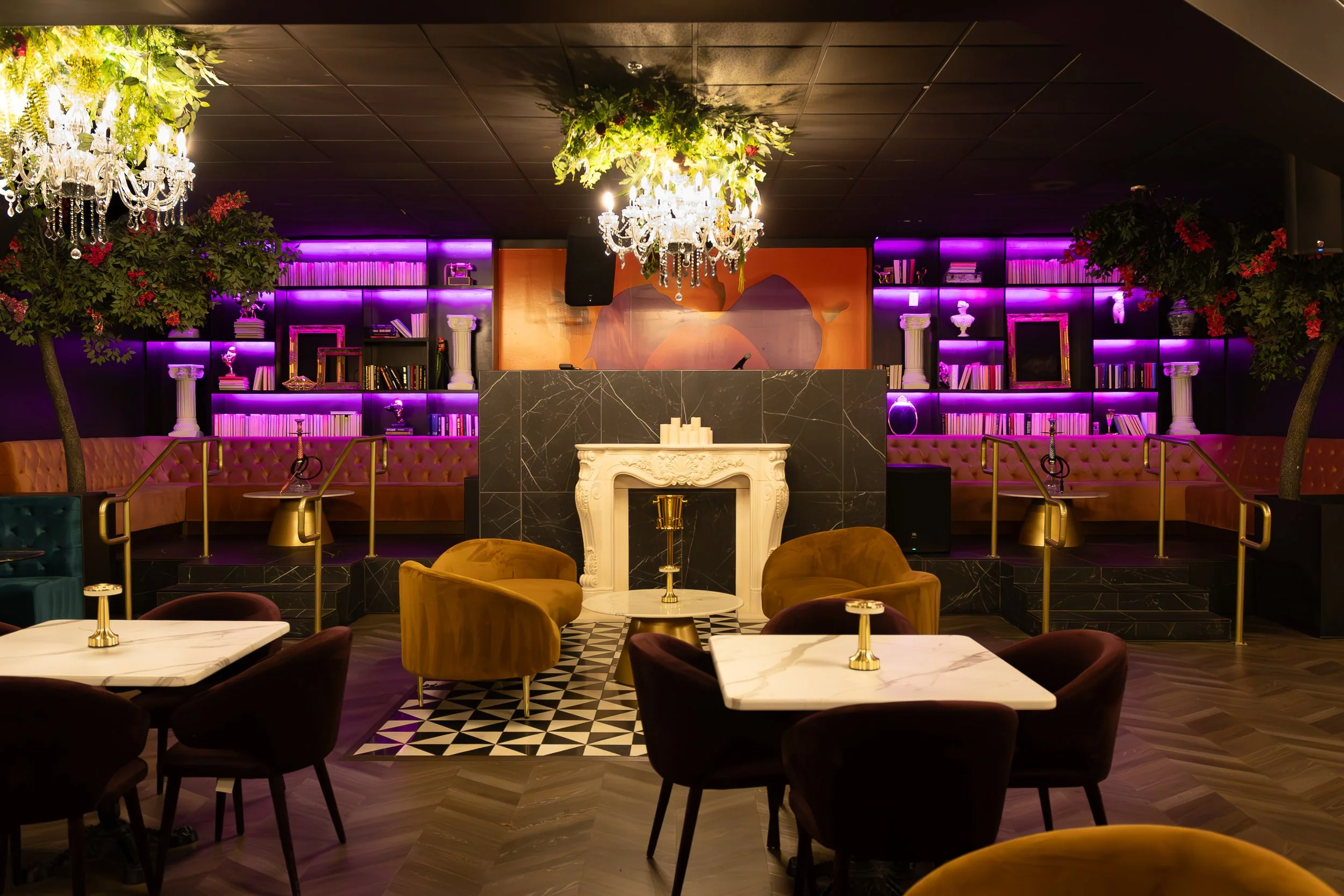 Interior view of a stylish lounge or bar with purple lighting, plush seating, marble-topped tables, decorative chandeliers, and bookshelves with purple backlighting.