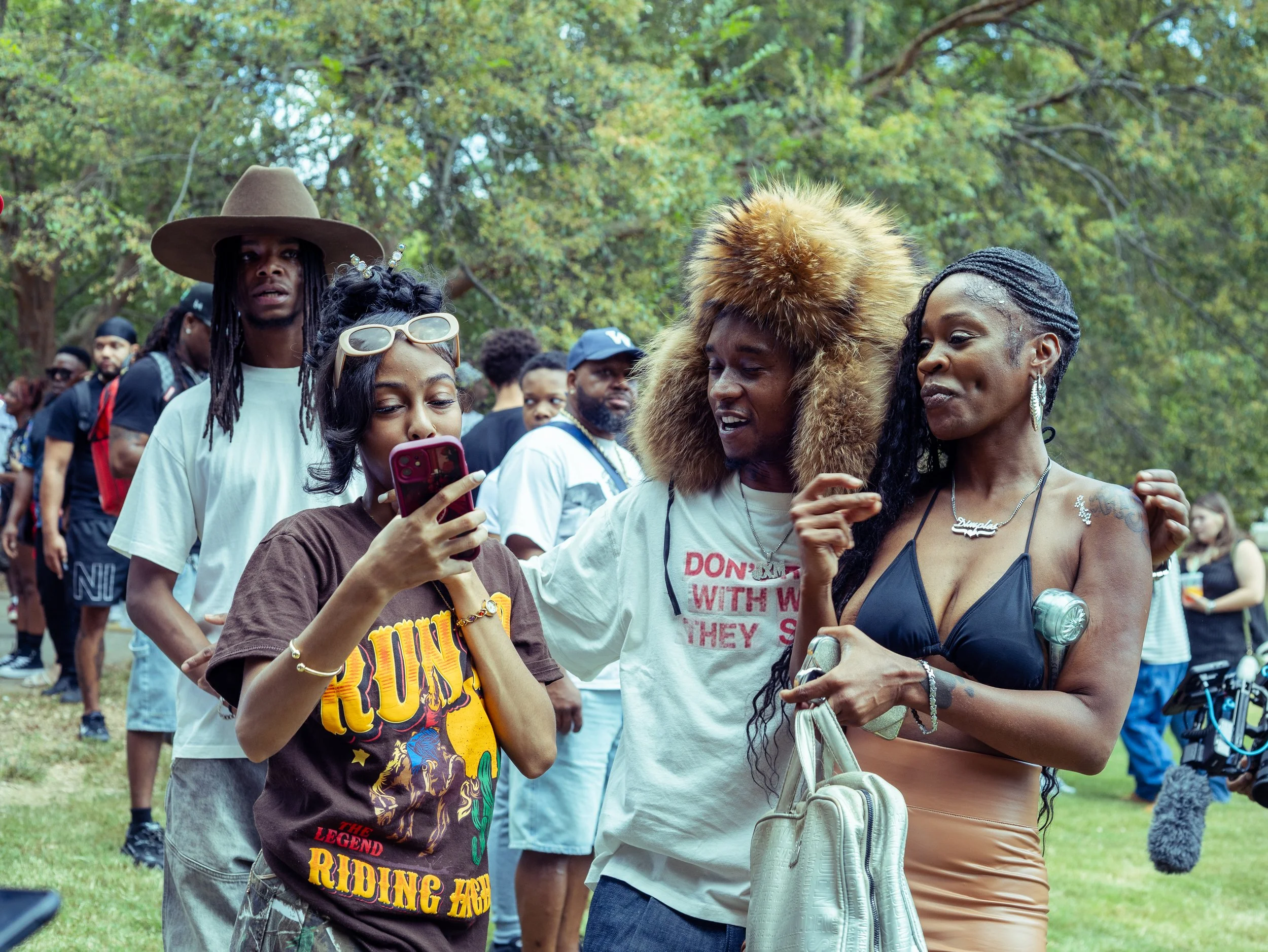 Group of diverse people socializing outdoors in a park with trees in the background, some taking photos or videos, dressed casually.