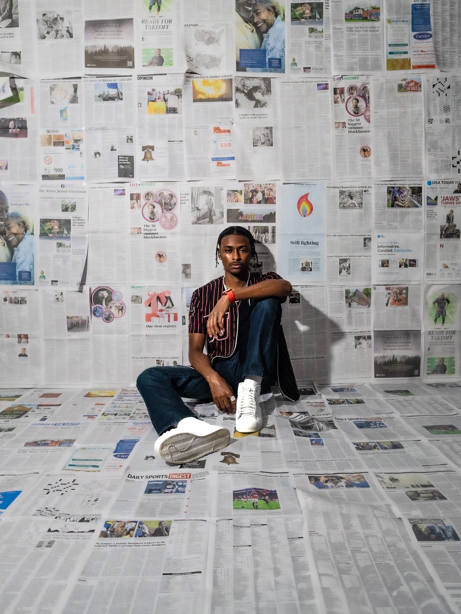 A young man with dark skin, wearing a black and red striped shirt, jeans, and white sneakers, sitting on the floor against a wall covered entirely with newspapers, with newspapers also spread across the floor in front of him.