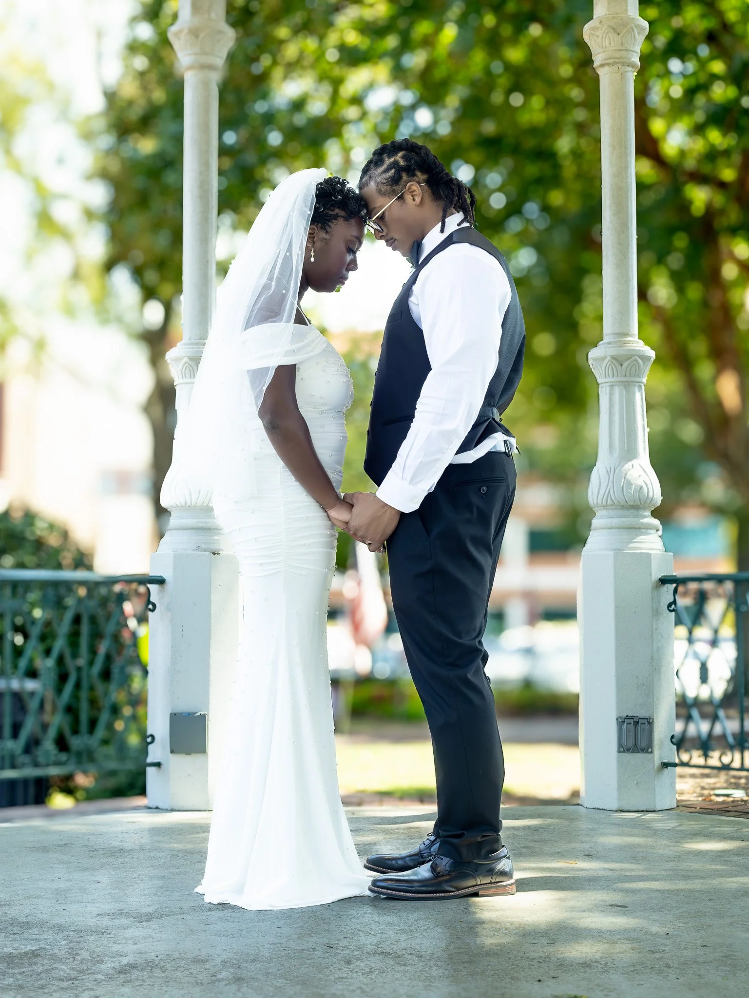 A couple in wedding attire holding hands and bowing their heads in prayer or reflection in a park with trees and a gazebo structure.