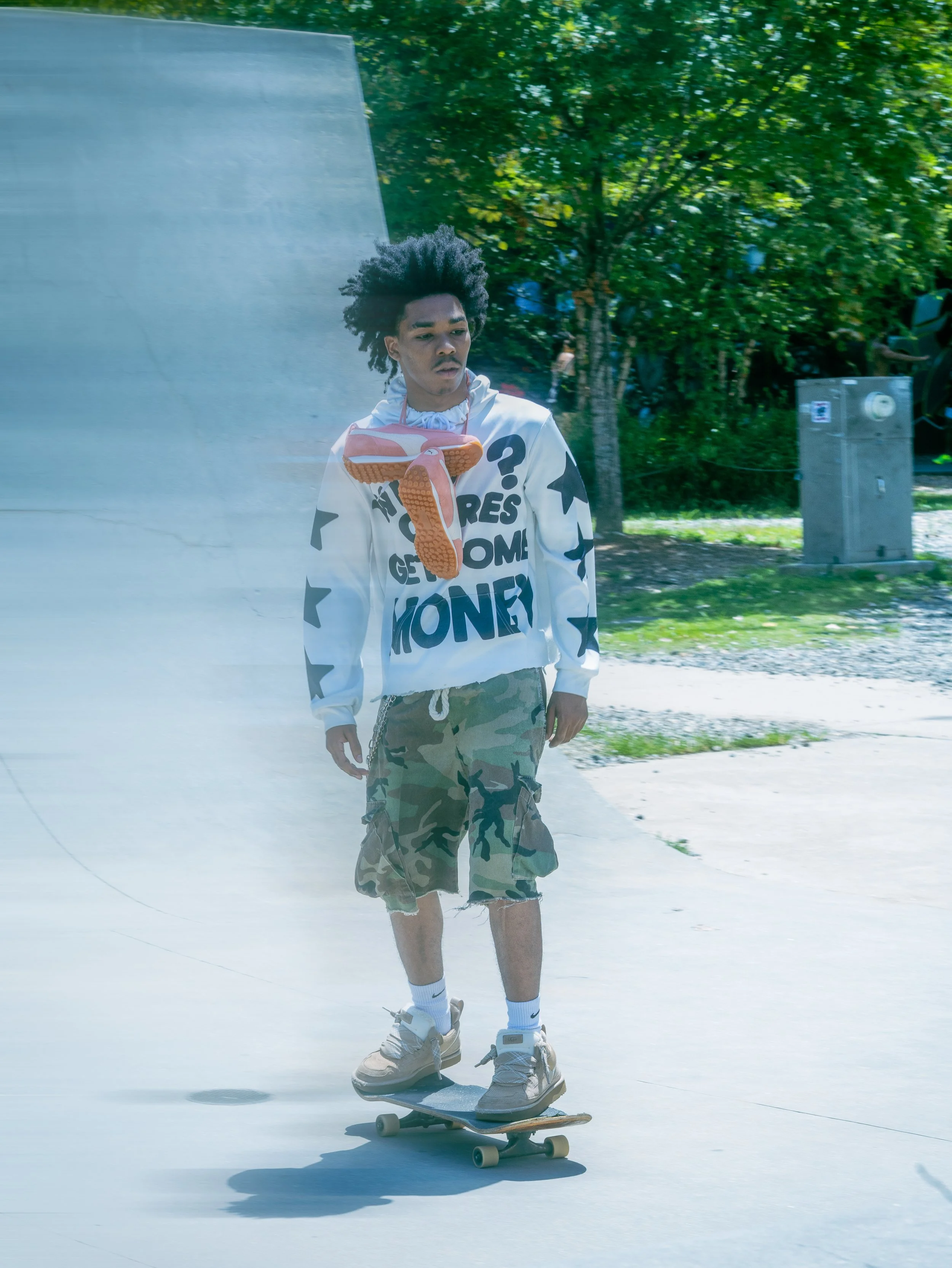 Young man skateboarding outdoors in a skatepark, wearing a white hoodie, camo shorts, and sneakers, with a shoe tied around his neck.