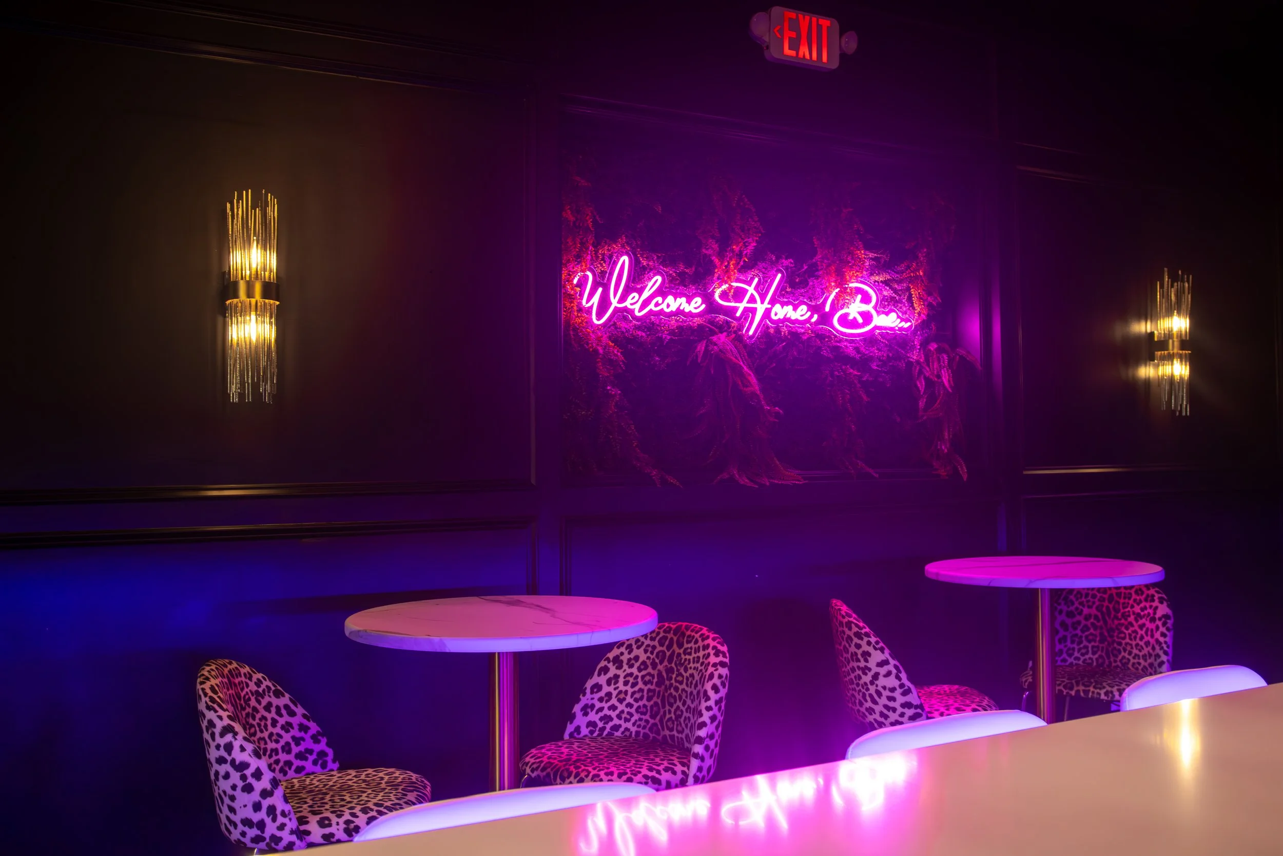 A dimly lit lounge area with leopard print chairs around small round marble tables, neon pink sign on the wall reading 'Welcome Home, Babe', and gold wall sconces with vertical light fixtures. An illuminated red exit sign is visible at the top.