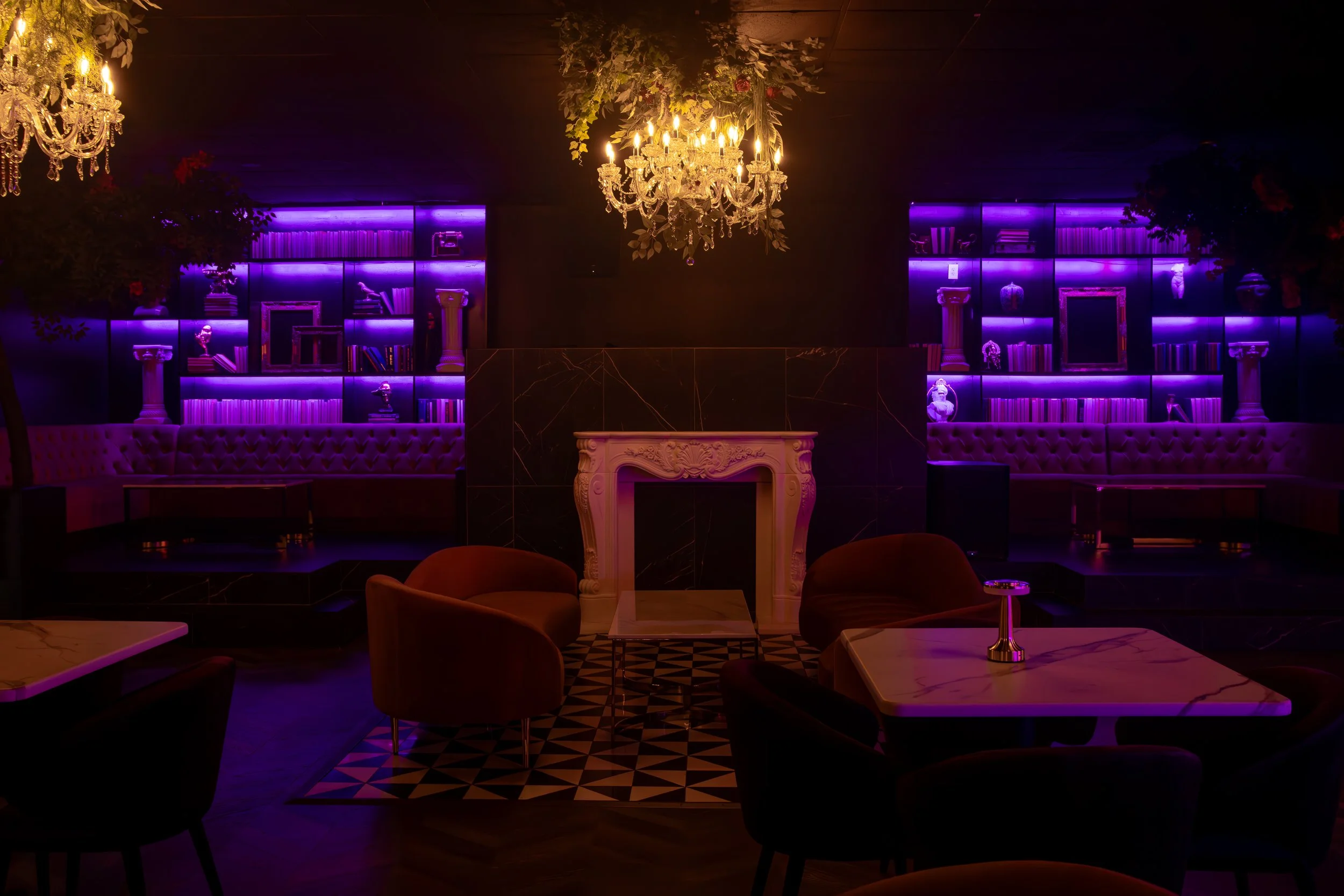 A dimly lit lounge with purple neon lights illuminating bookshelves, a white ornate fireplace, and plush seating. Two chandeliers hang from the ceiling, casting warm light.