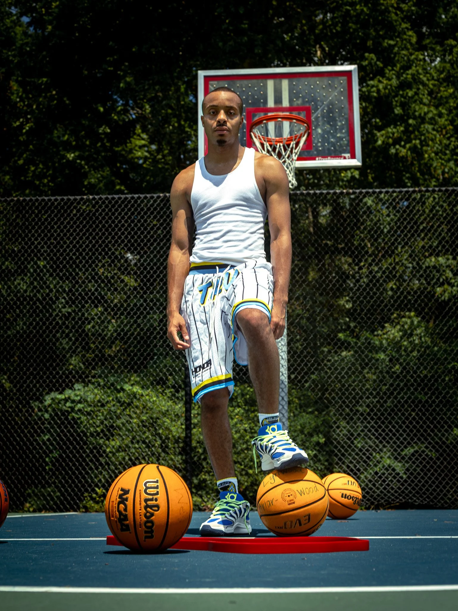 A young man standing on a basketball court with a basketball dribbling under his foot, surrounded by three additional basketballs, with a basketball hoop in the background.