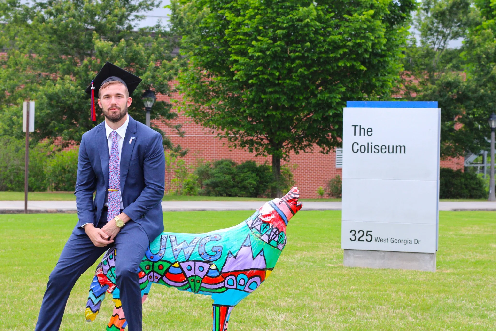 A young man in a blue suit, graduation cap, and colorful tie sitting on a painted horse sculpture outside. Behind him is a sign that reads 'The Coliseum, 325 West Georgia Dr,' with green trees and a brick building in the background.