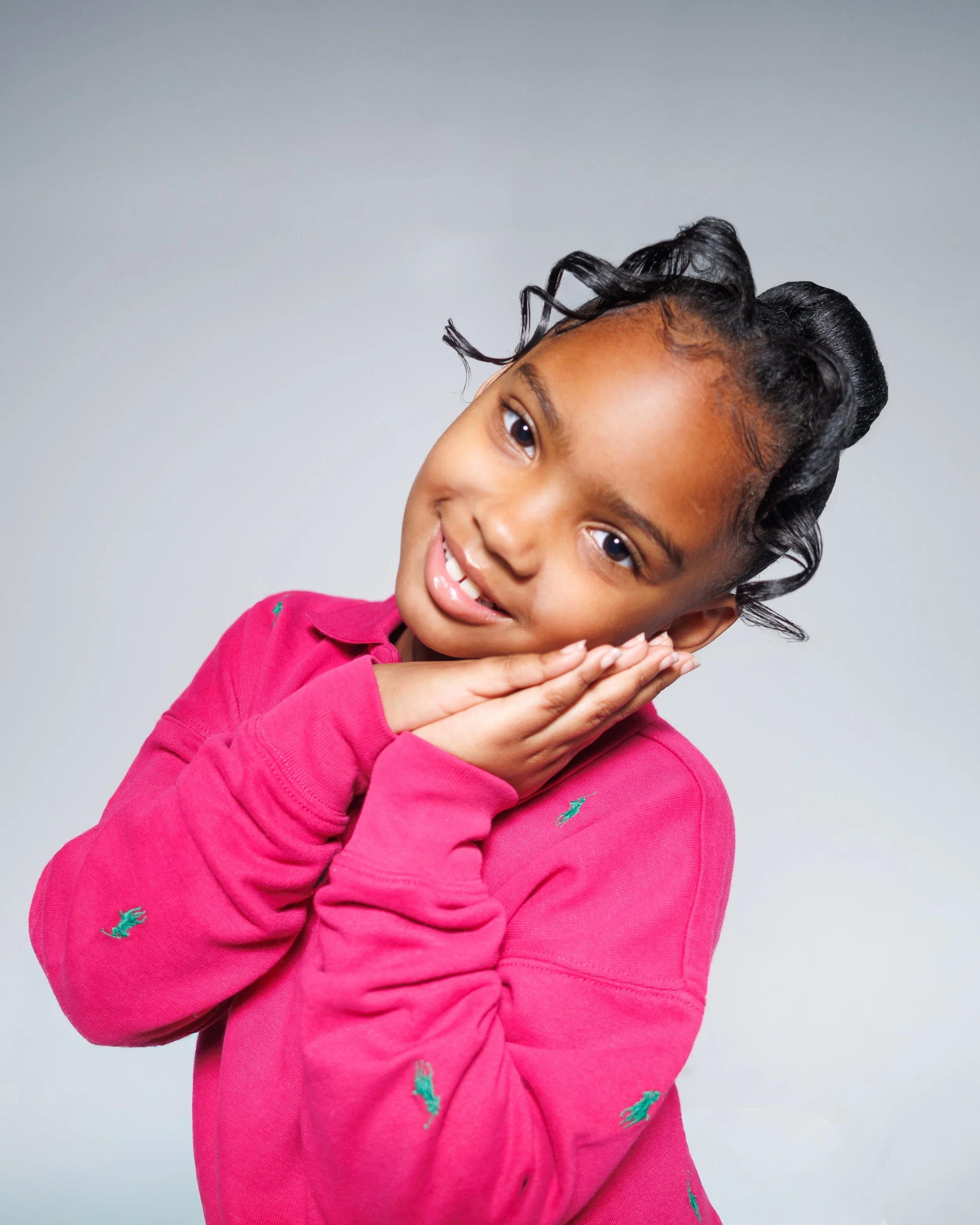 Young girl with curly hair styled in rollers, wearing a pink sweatshirt, smiling and resting her face on her hands against a gray background.