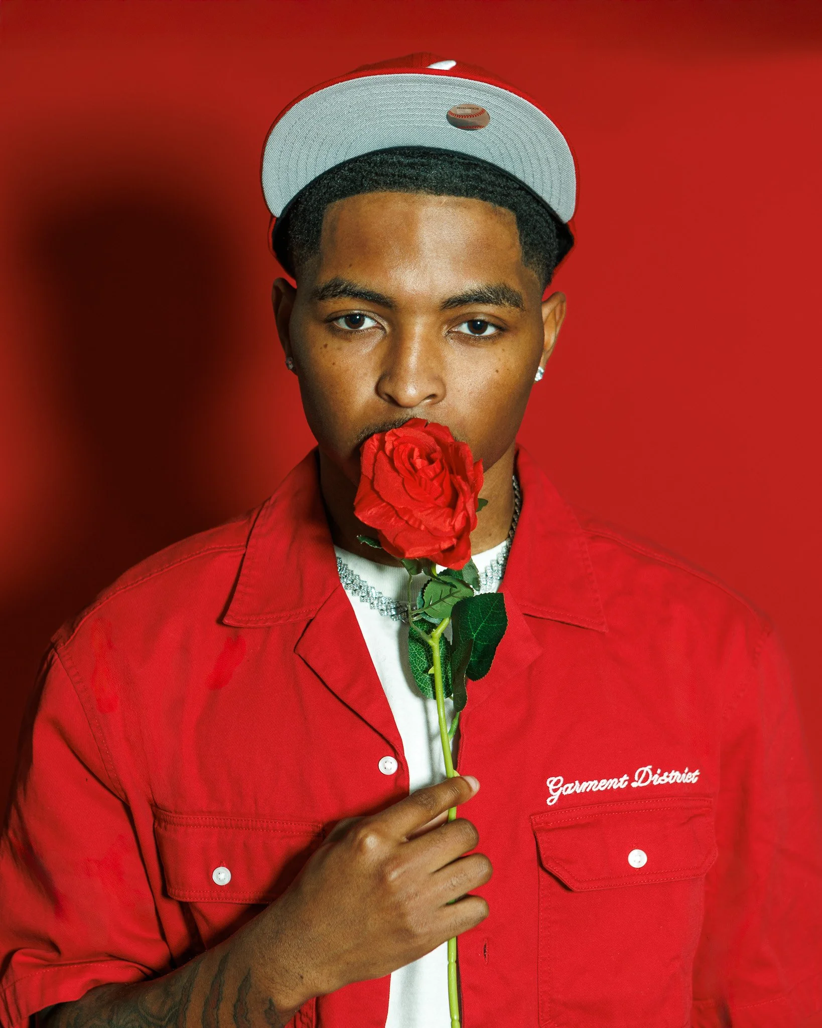 Young man with short hair, wearing a white cap with a red logo, a red jacket with 'Garment District' embroidered on it, holding a red rose near his lips, against a red background.