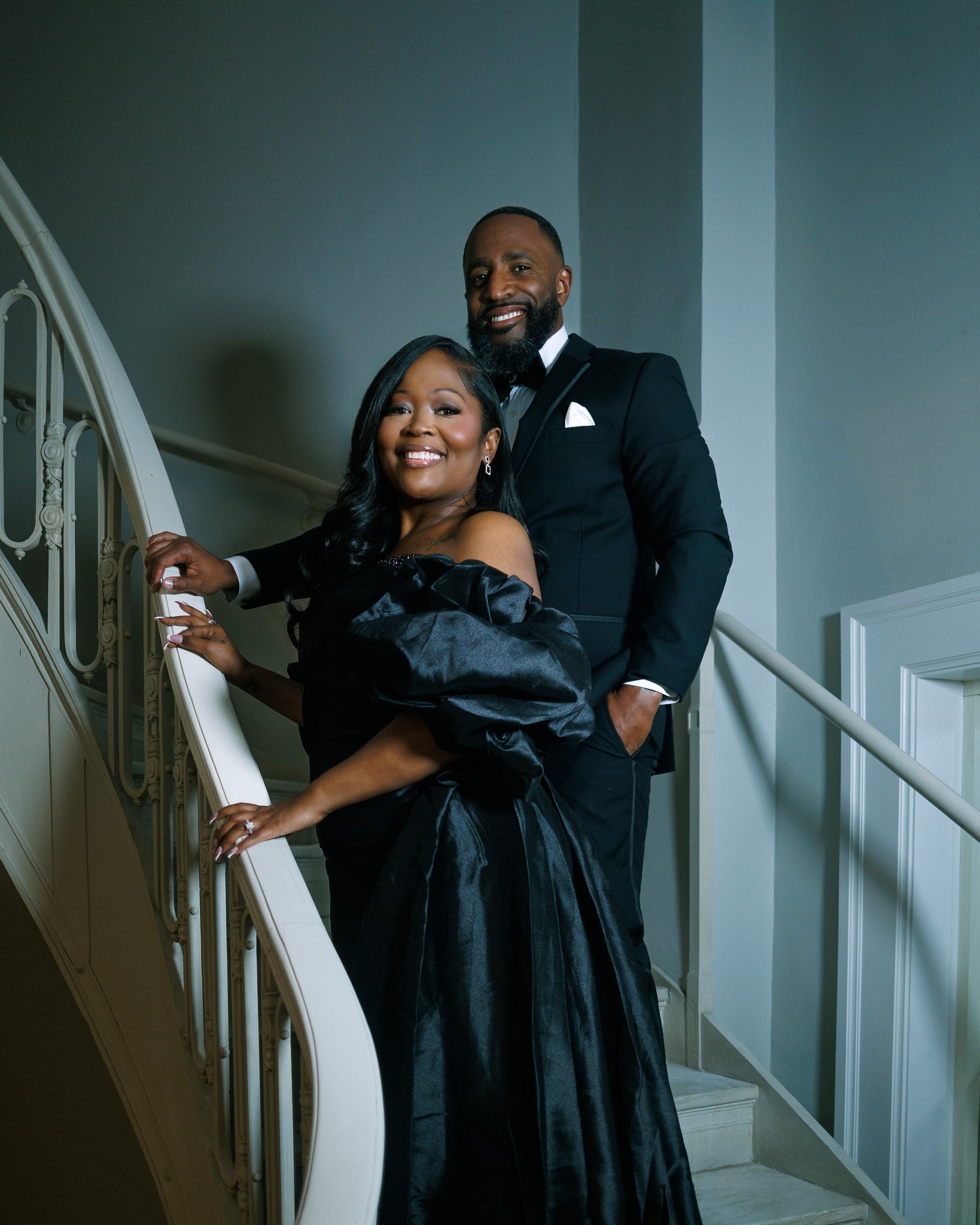 A couple dressed in formal attire standing on a staircase, smiling for the camera. The woman is leaning on the railing, wearing an off-the-shoulder black evening gown with puffy sleeves, and the man is standing behind her in a black tuxedo with a bow