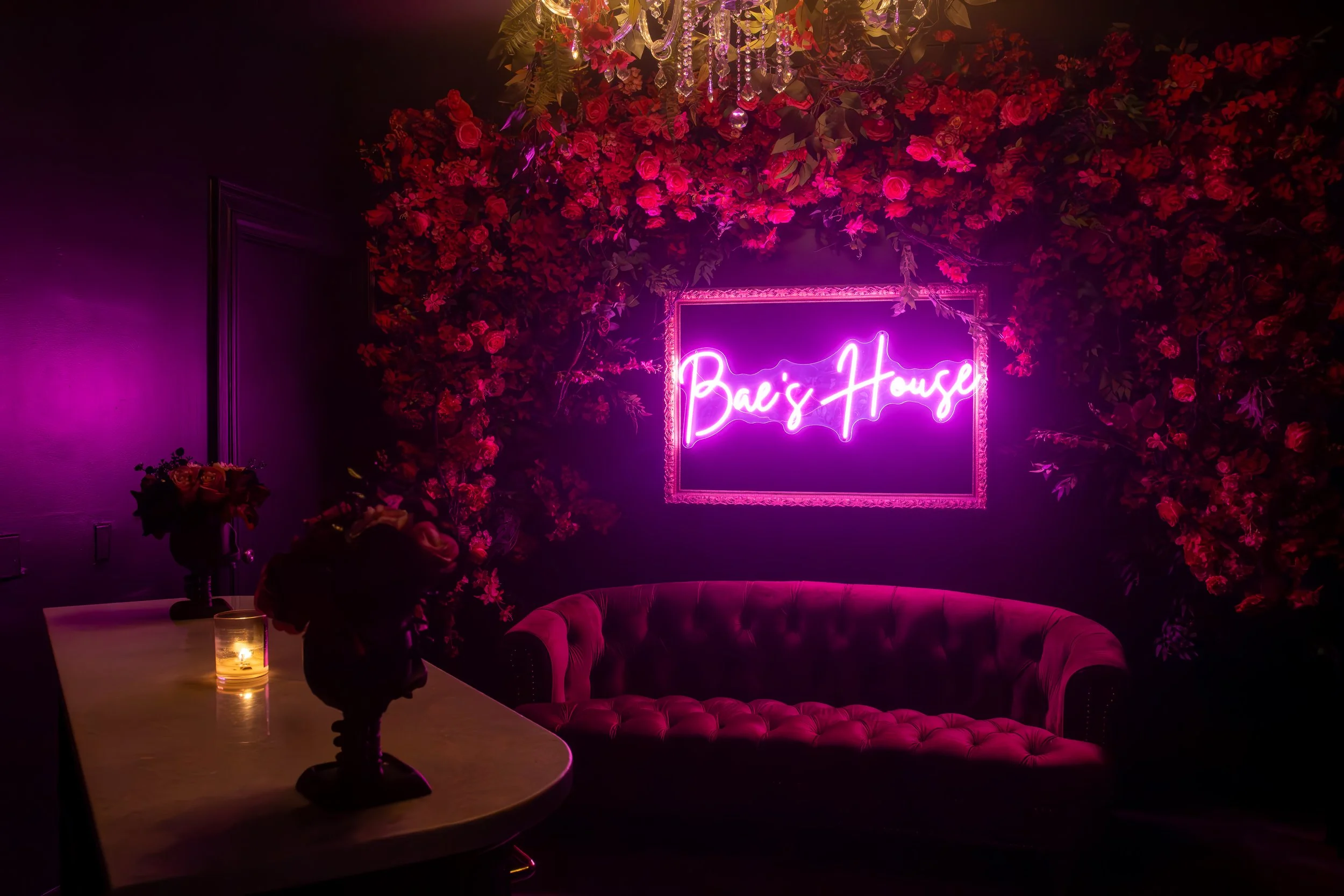 A dimly lit, elegant lounge area with pink and purple lighting. A neon sign reads 'Bai's House,' surrounded by a lush wall of pink flowers. There is a pink velvet sofa beneath the sign and a table on the left with flower arrangements and a lit candle