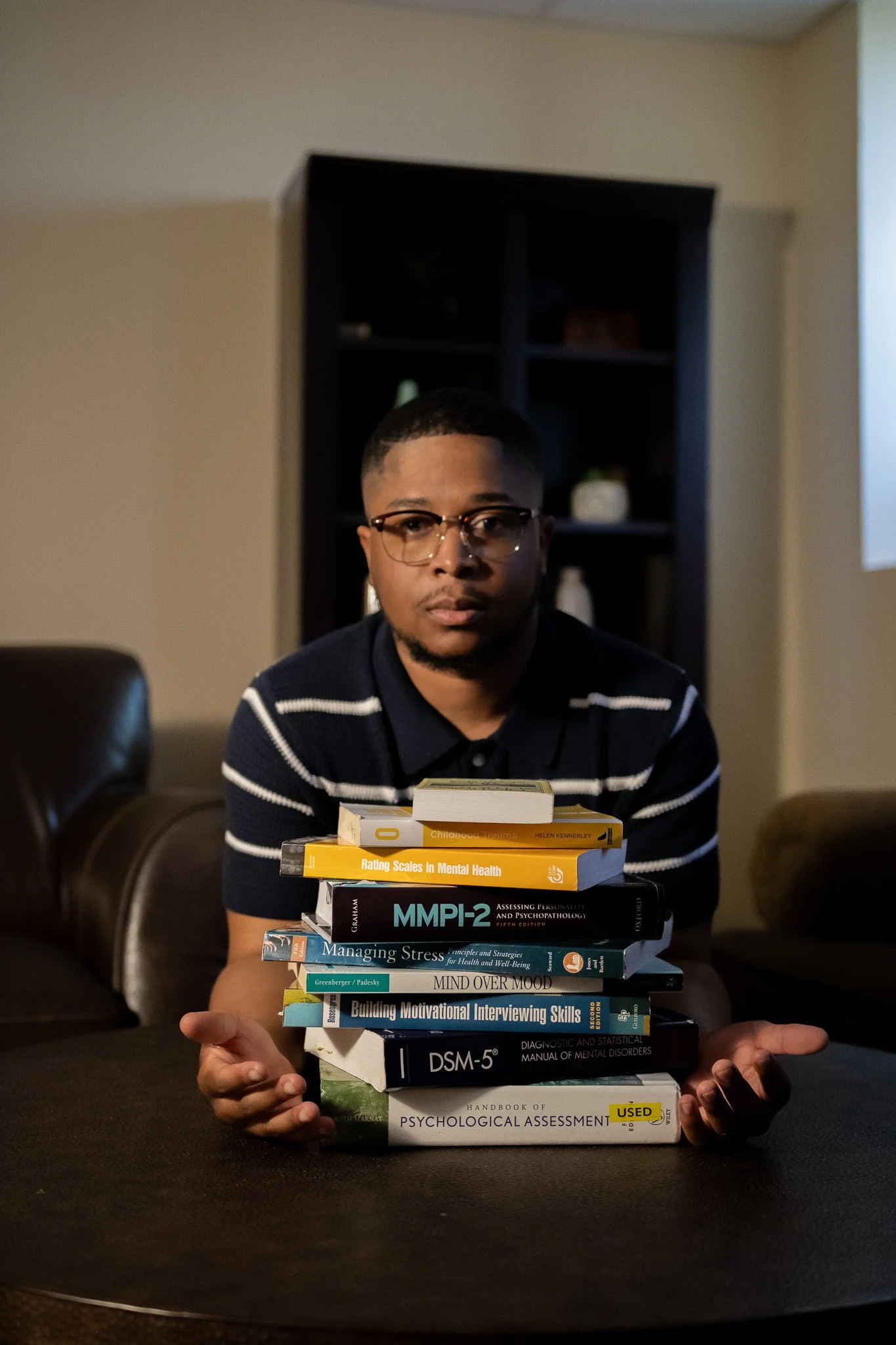 A man with glasses and a striped shirt sitting at a table, holding a stack of mental health and psychology books.