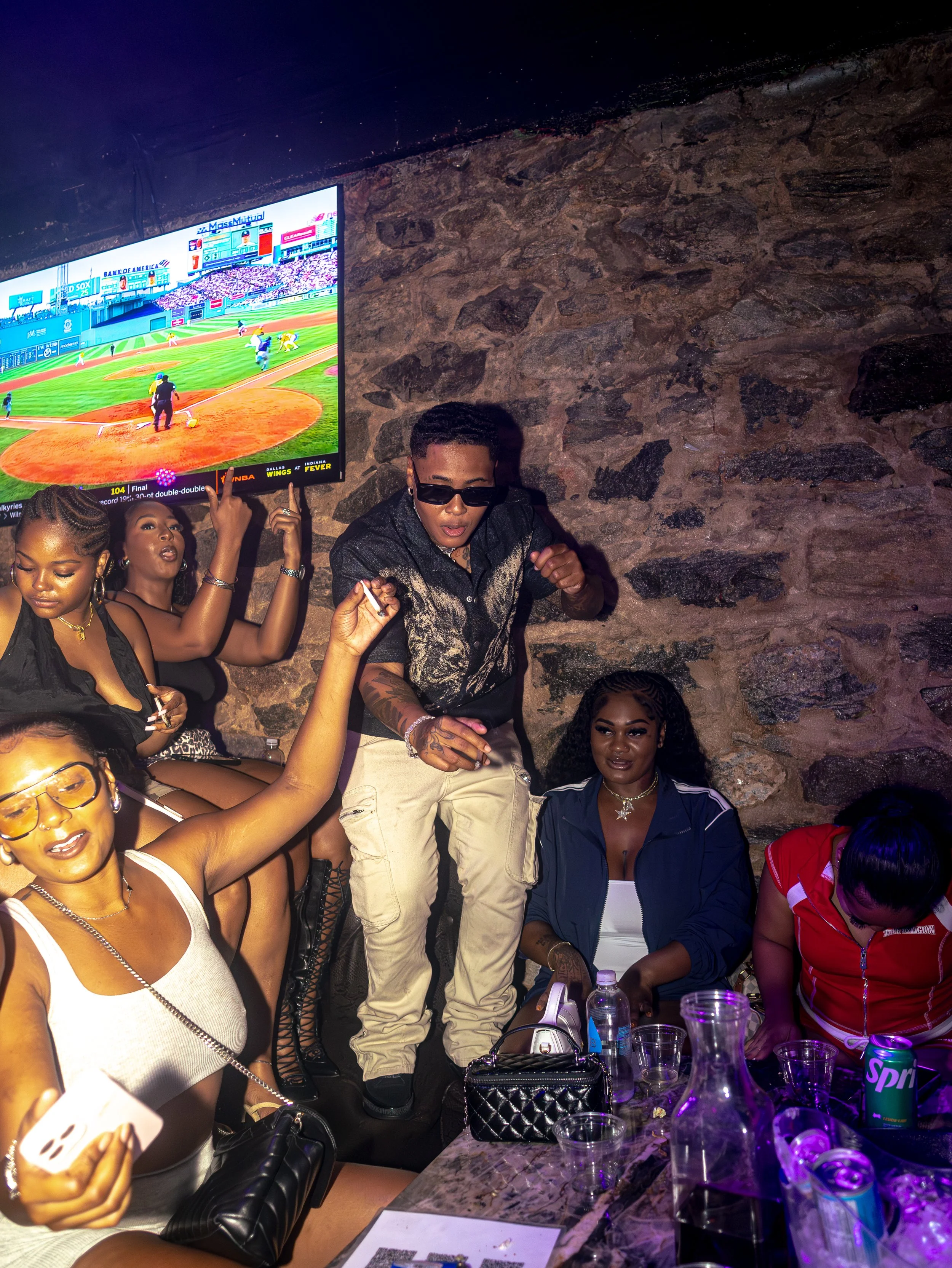 Group of young women and a man at party or gathering, some sitting, some dancing, with drinks and snacks on table, stone wall background, TV screen showing baseball game.