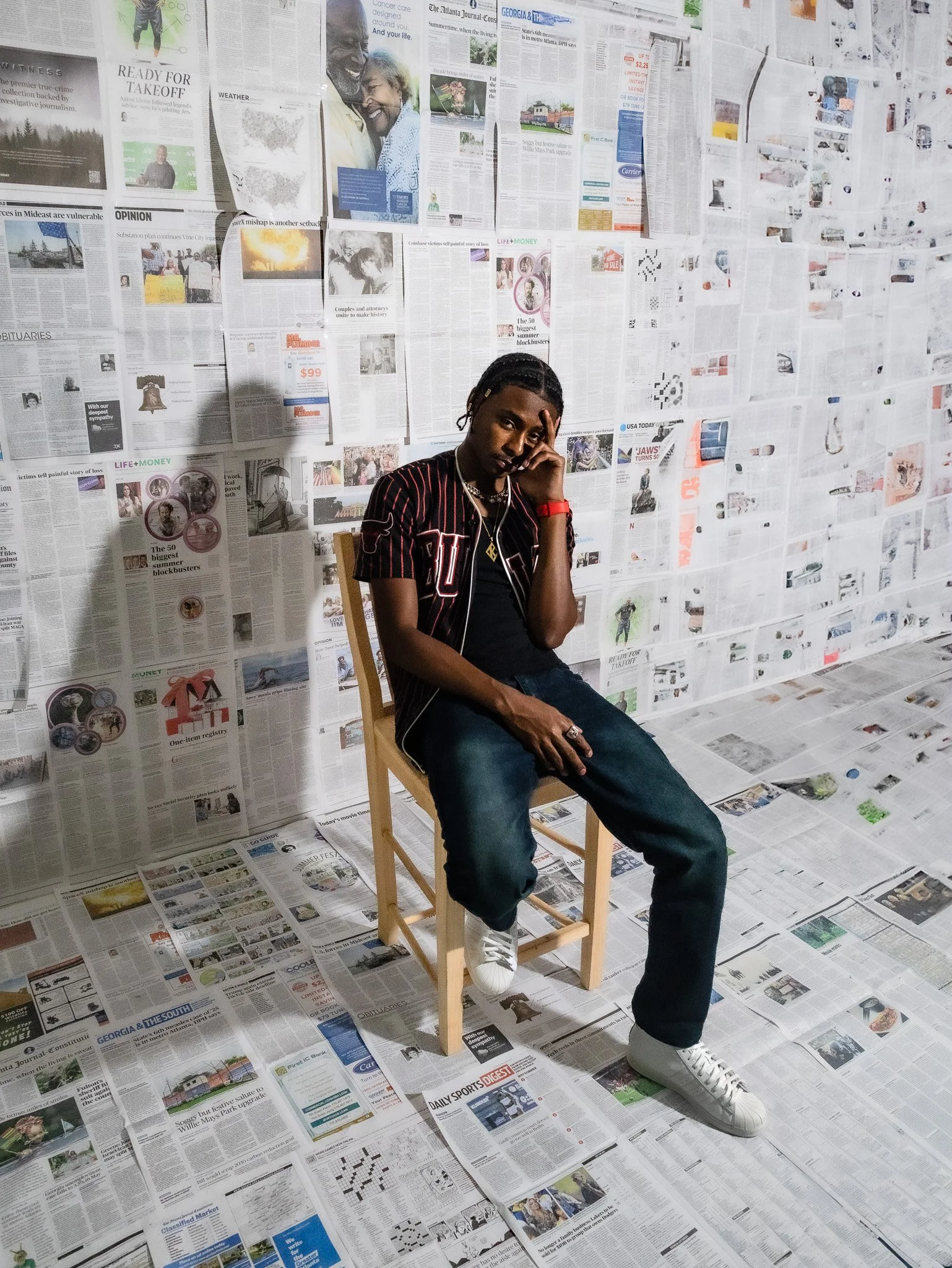A man sitting on a wooden chair in a room with walls and floor covered intentionally with newspapers, making a backdrop and floor covering.