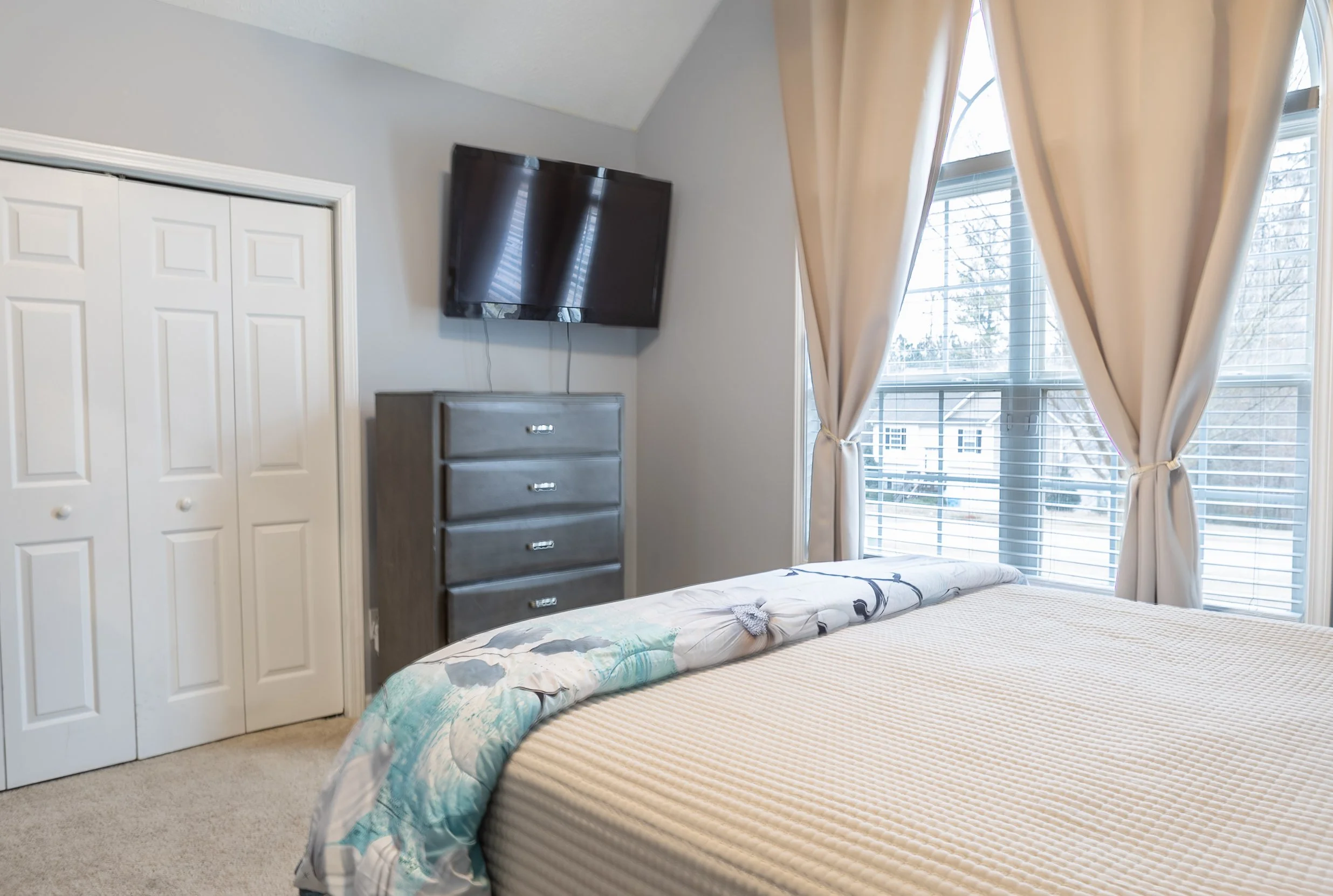 A bedroom with a bed, window with curtains, a dresser, and a wall-mounted TV.