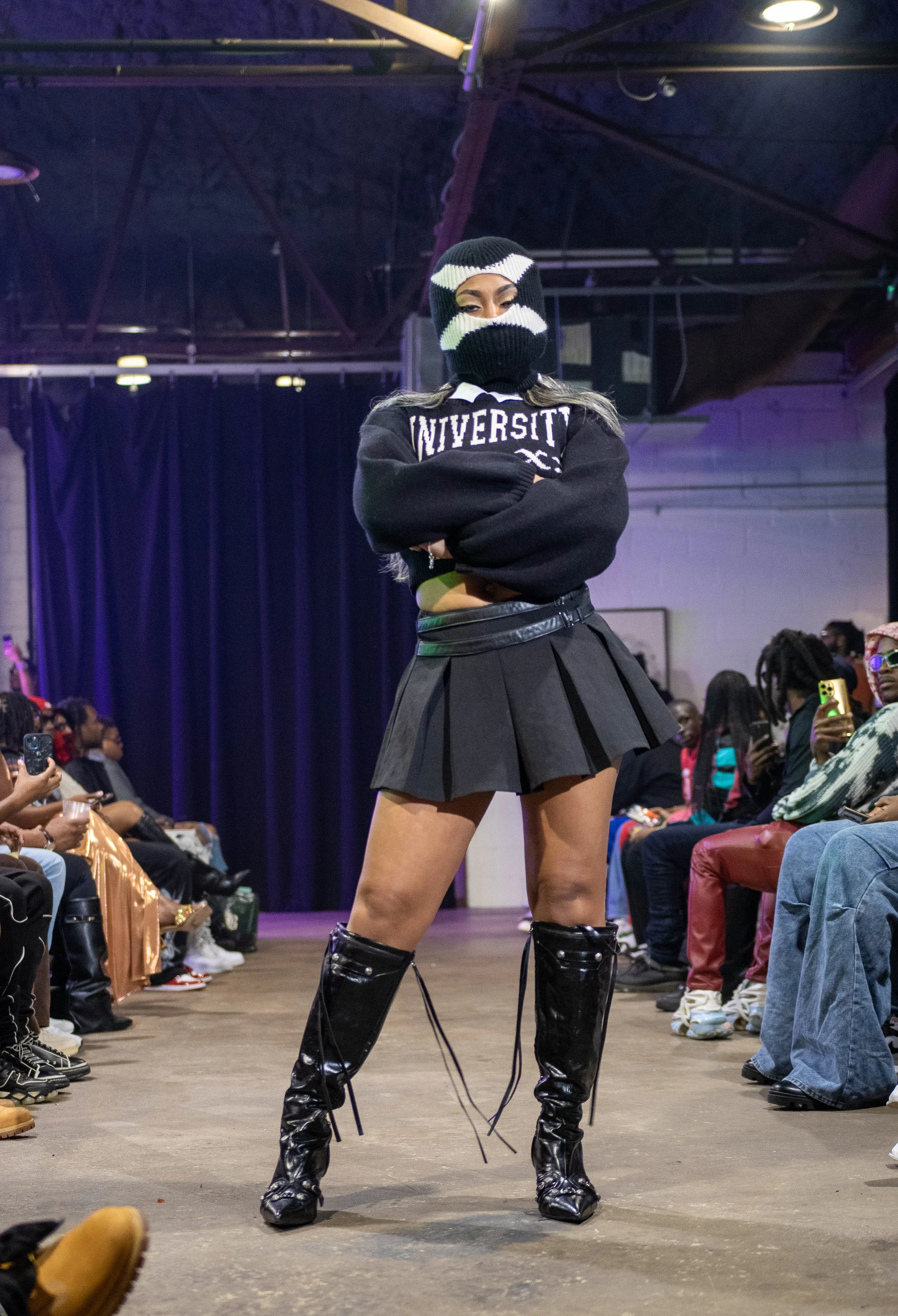 A woman walking down a runway during a fashion show, wearing a black sweater with 'UNIVERSITY' printed on it, a black pleated skirt, shiny black knee-high boots with laces, and a black and white ski mask covering her face, with the audience seated on