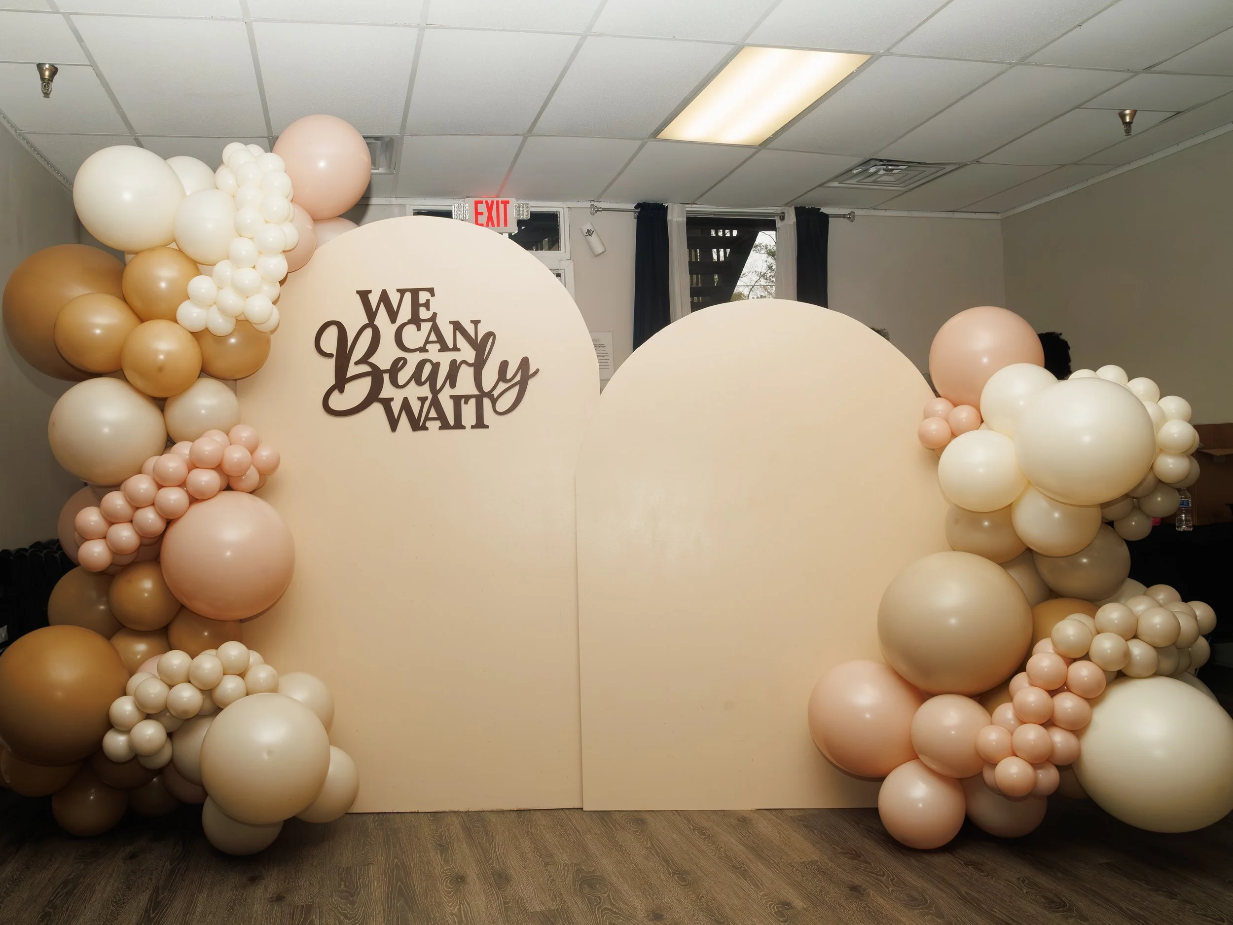 A decorated backdrop with balloons in shades of white, cream, blush, and gold, with a round sign that says 'We Can Bearly Wait' at an event or celebration.