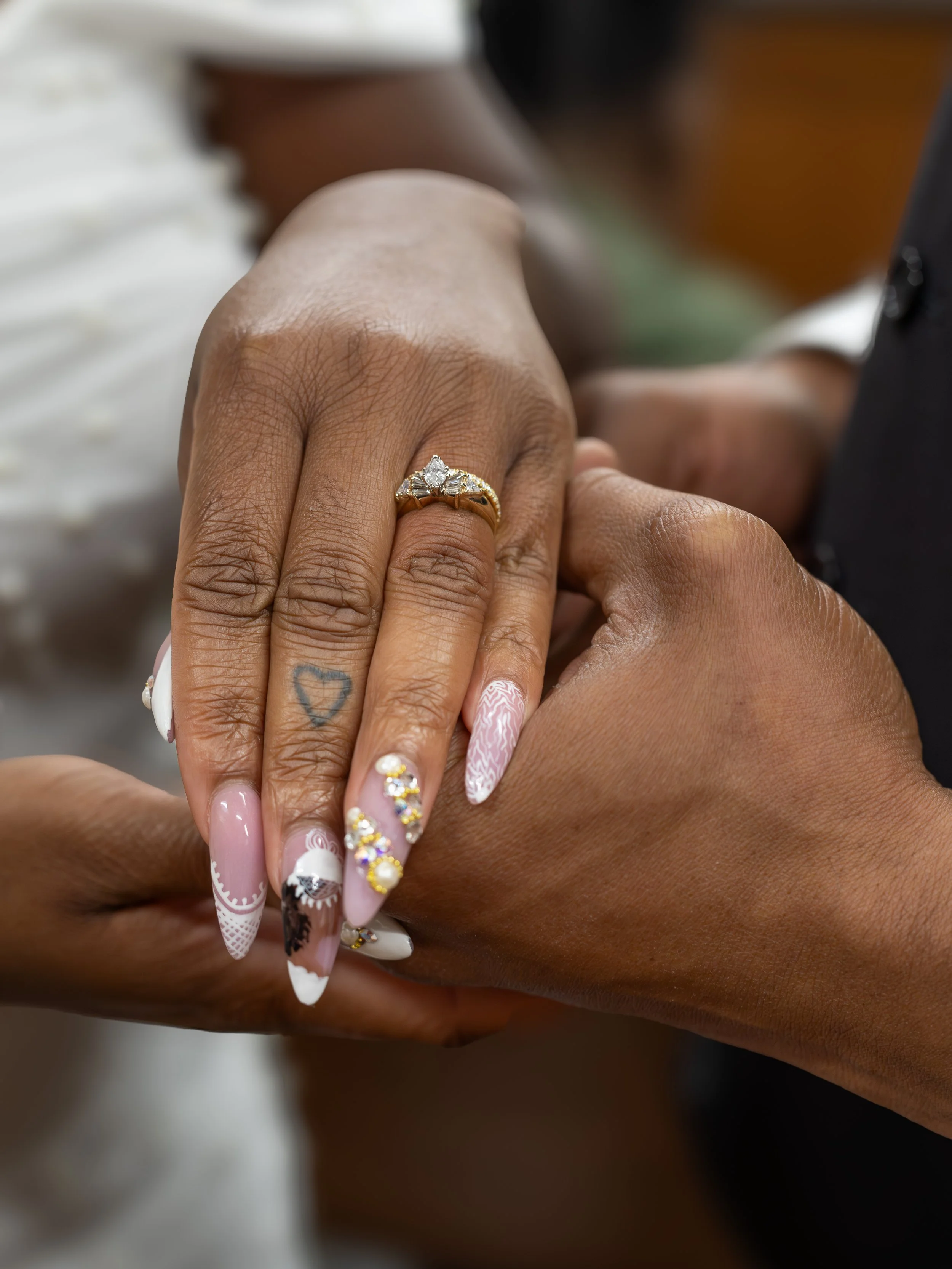 Close-up of two hands holding each other, with the focus on one hand wearing a gold ring with a diamond. Both hands have manicured nails with intricate nail art and embellishments. One hand has a small heart tattoo on the middle finger.