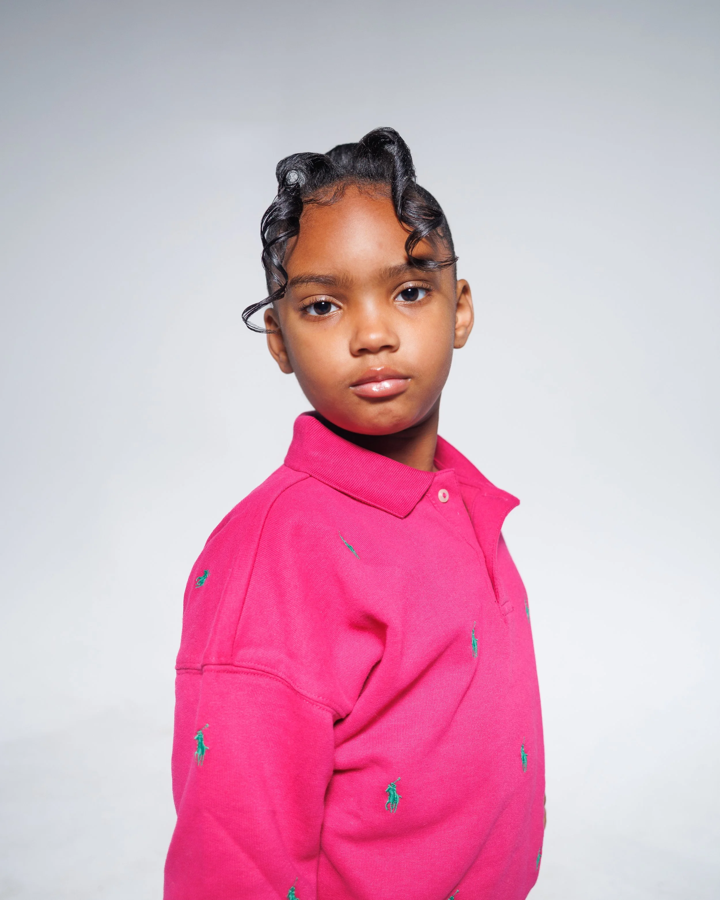 A young girl with curly hair styled in rings, wearing a pink polo shirt with small green embroidered logos, looking directly at the camera against a plain light grey background.