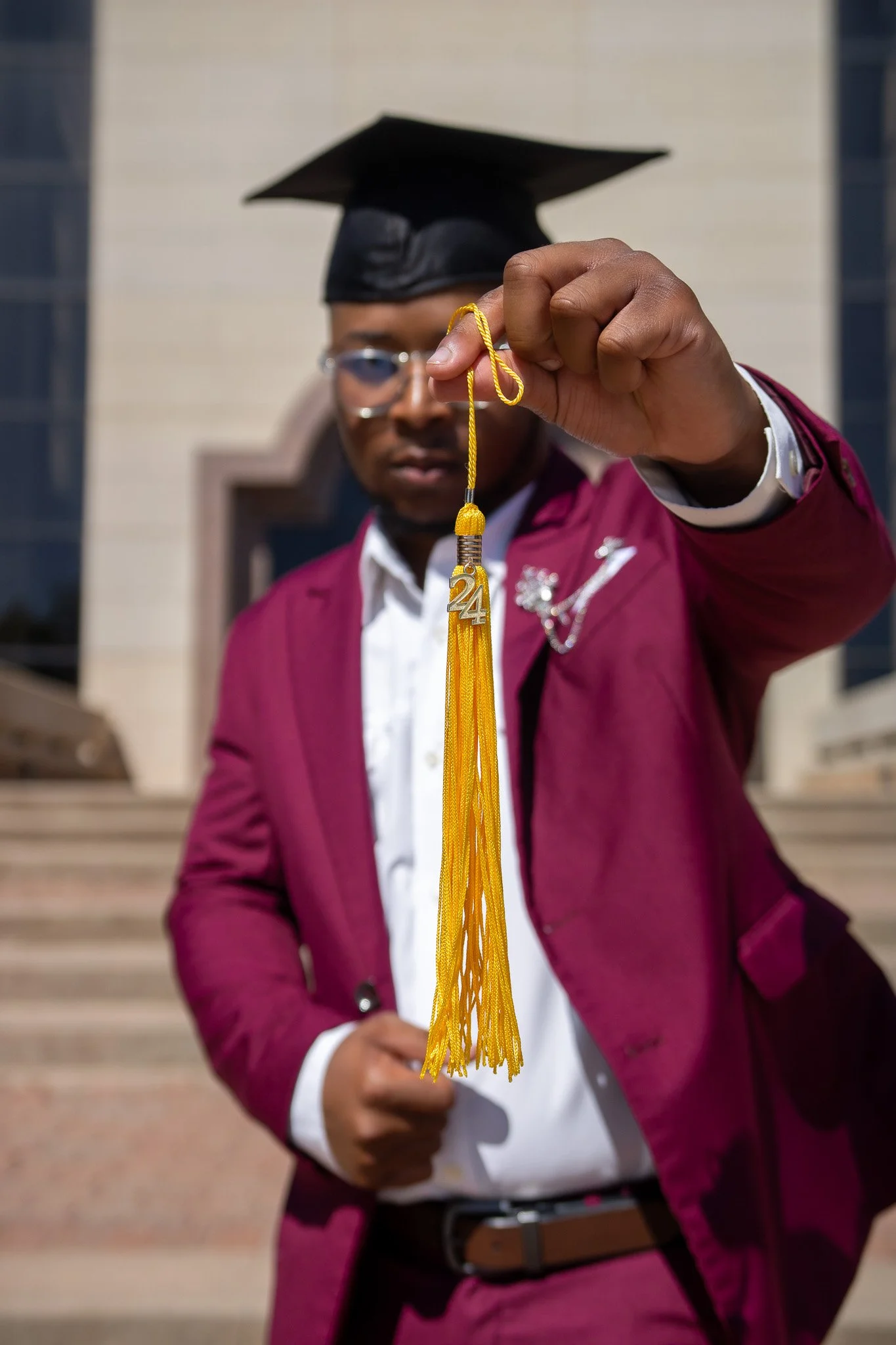Man in maroon suit and cap holding a yellow tassel with a '24' charm in front of a building steps.