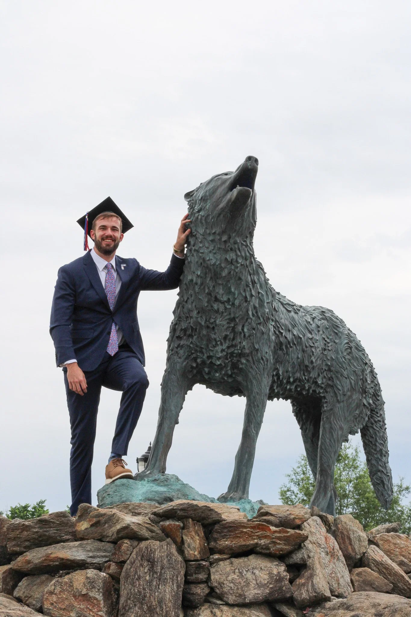 A man wearing a graduation cap and suit standing next to a large wolf statue, touching its head with one hand, on a rocky base, outdoors with a cloudy sky.