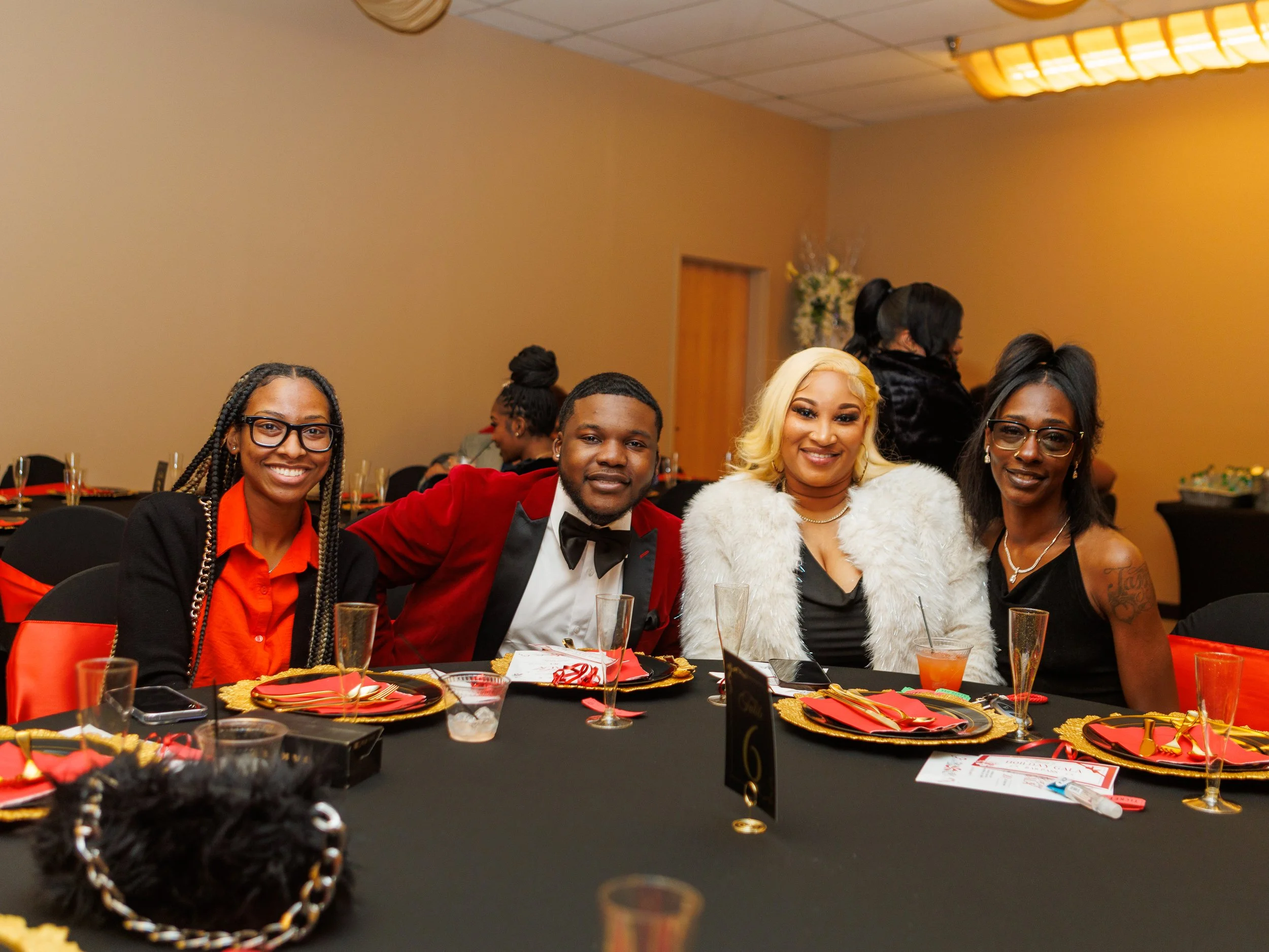 Four people sitting at a decorated banquet table, smiling and dressed in formal attire, at an indoor event or celebration.