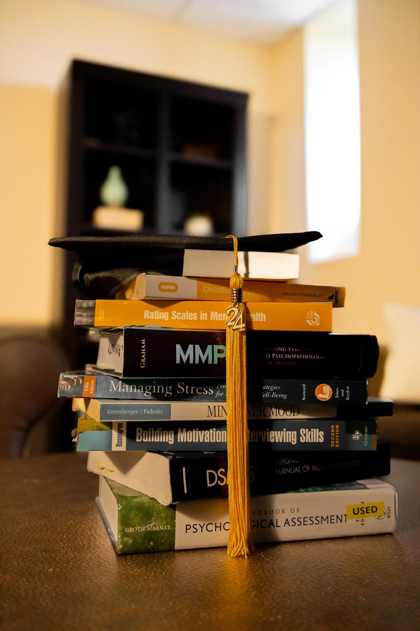 Stack of psychology and mental health books topped with a graduation cap and tassel in a cozy room with a bookshelf and a window in the background.