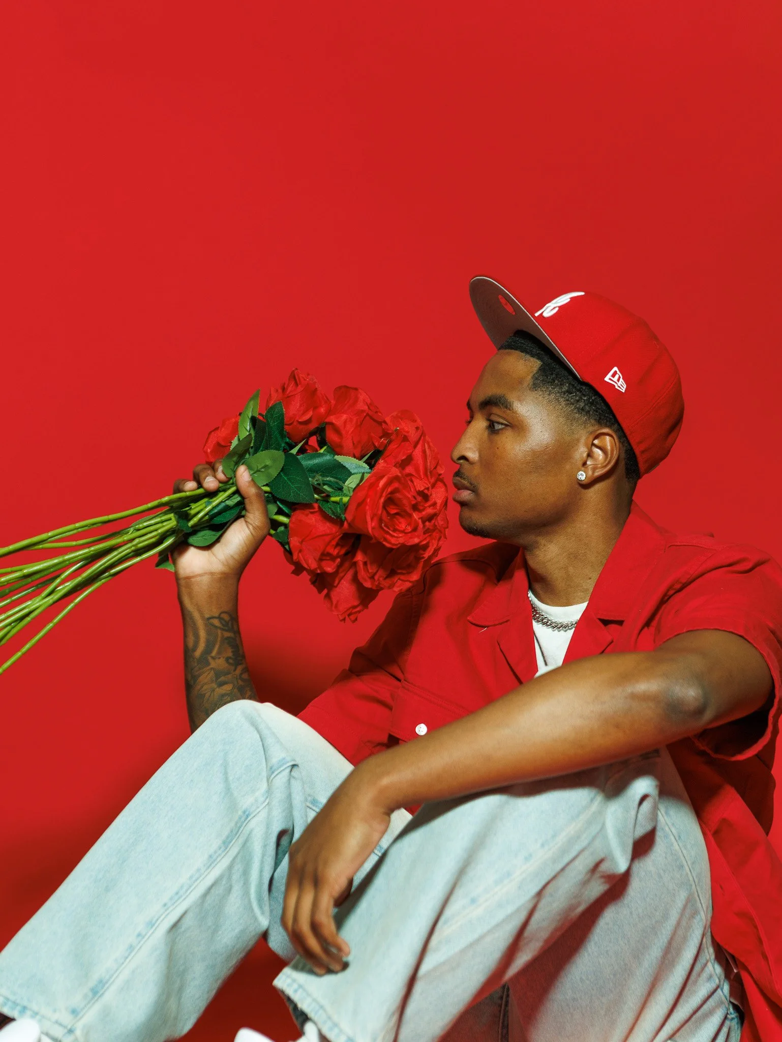 A young man in a red shirt, gray cap, and jewelry sits against a red backdrop, holding a bouquet of red roses close to his face.