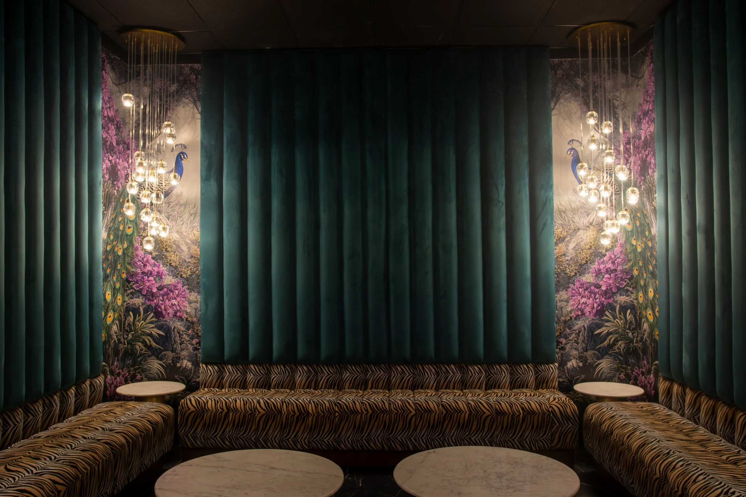 Luxurious lounge with dark green curtains, zebra-print seating, and floral wallpaper featuring pink and purple flowers and peacock images, illuminated by modern hanging light fixtures.