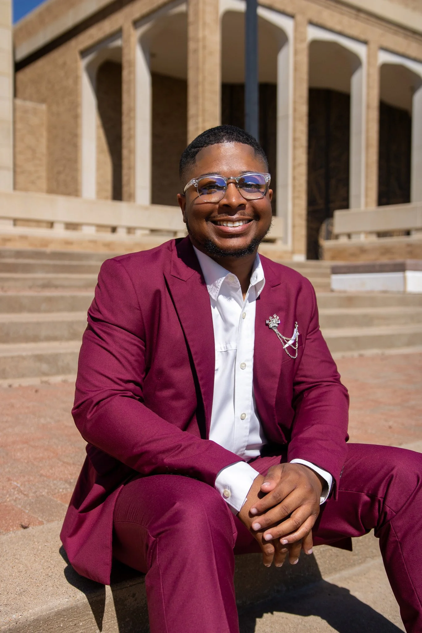 A man in a burgundy suit sitting on steps outside a building, smiling at the camera.