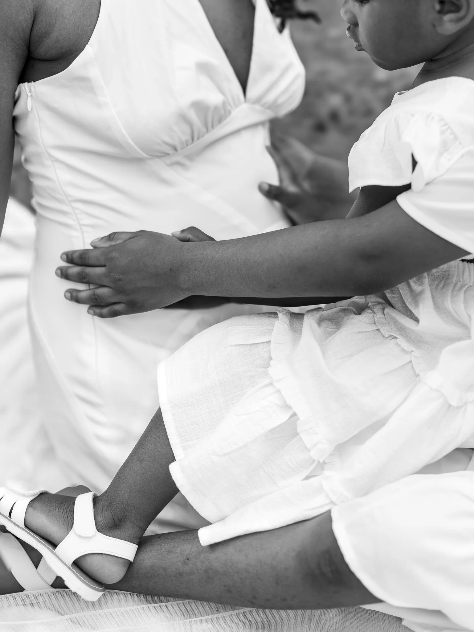 A woman with a pregnant belly holding her stomach while sitting next to a child, possibly her daughter, who is sitting on her lap. The woman and child are wearing light-colored clothing.