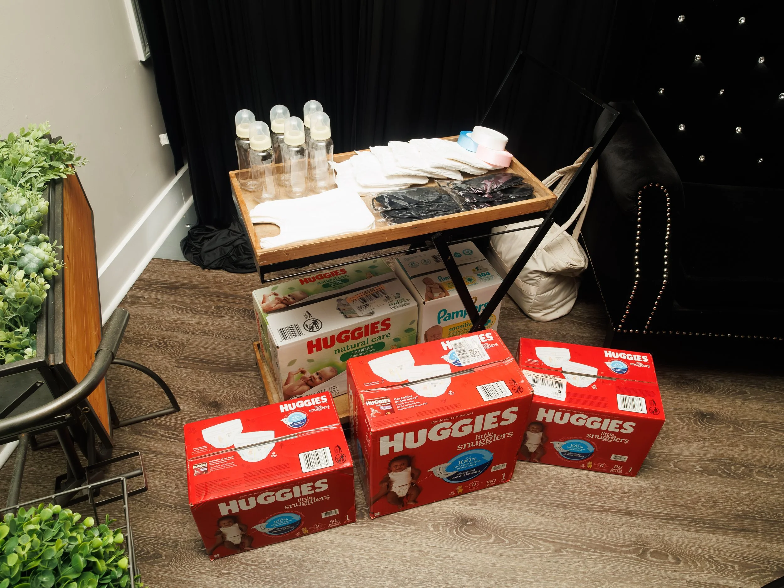 Stack of boxes of Huggies and Pampers diapers, a rolling table with baby bottles, wipes, and disposable gloves, and a black sofa in the background.