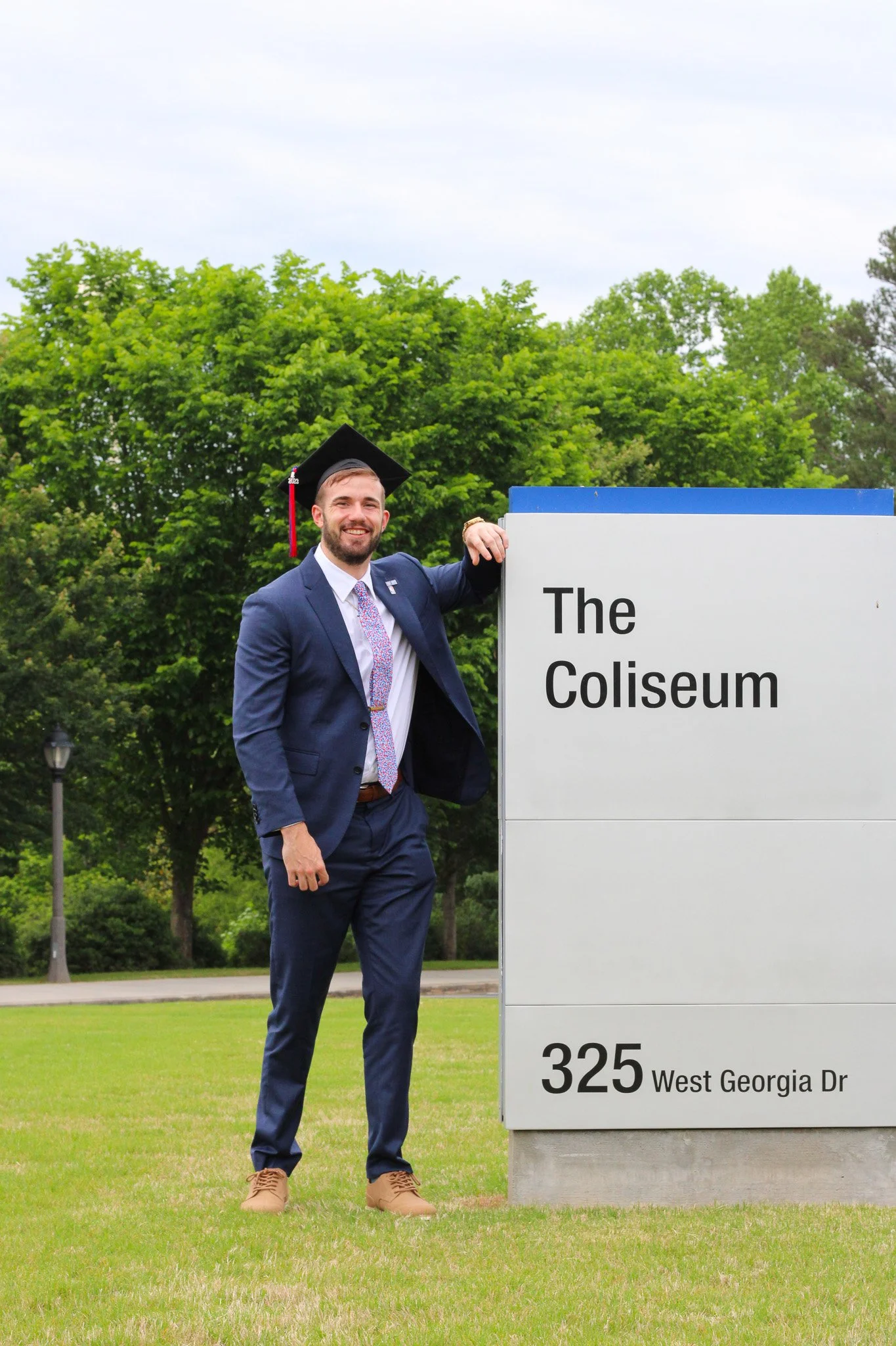 A man in a navy suit and graduation cap, smiling, standing next to a sign that reads 'The Coliseum' at 325 West Georgia Drive, with green trees and a grassy area in the background.