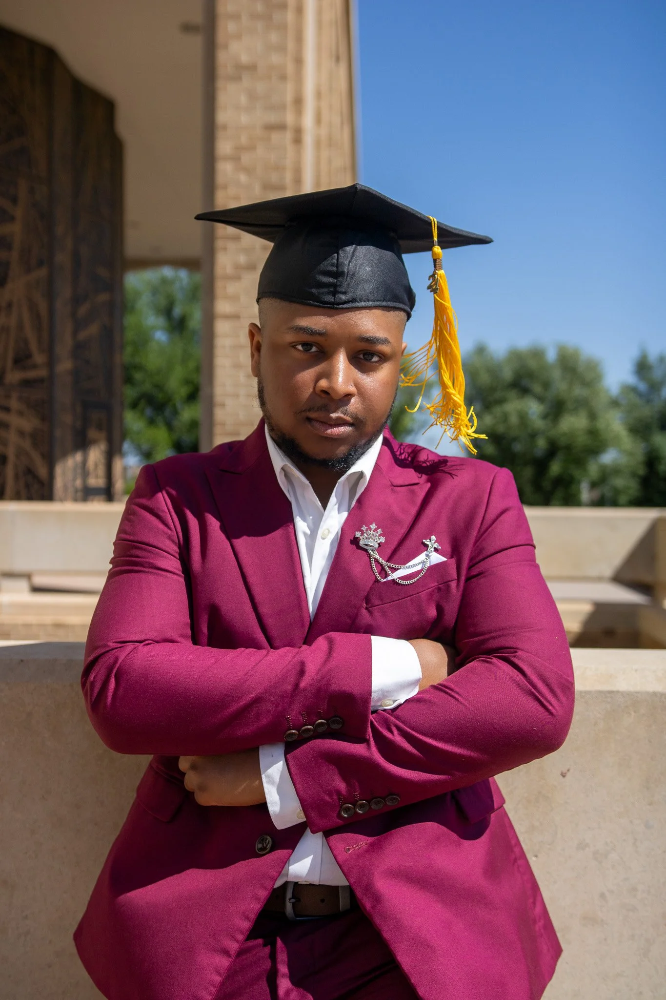 A young man in a maroon suit with a white shirt, and a black graduation cap with a yellow tassel, standing outdoors with arms crossed and looking at the camera.