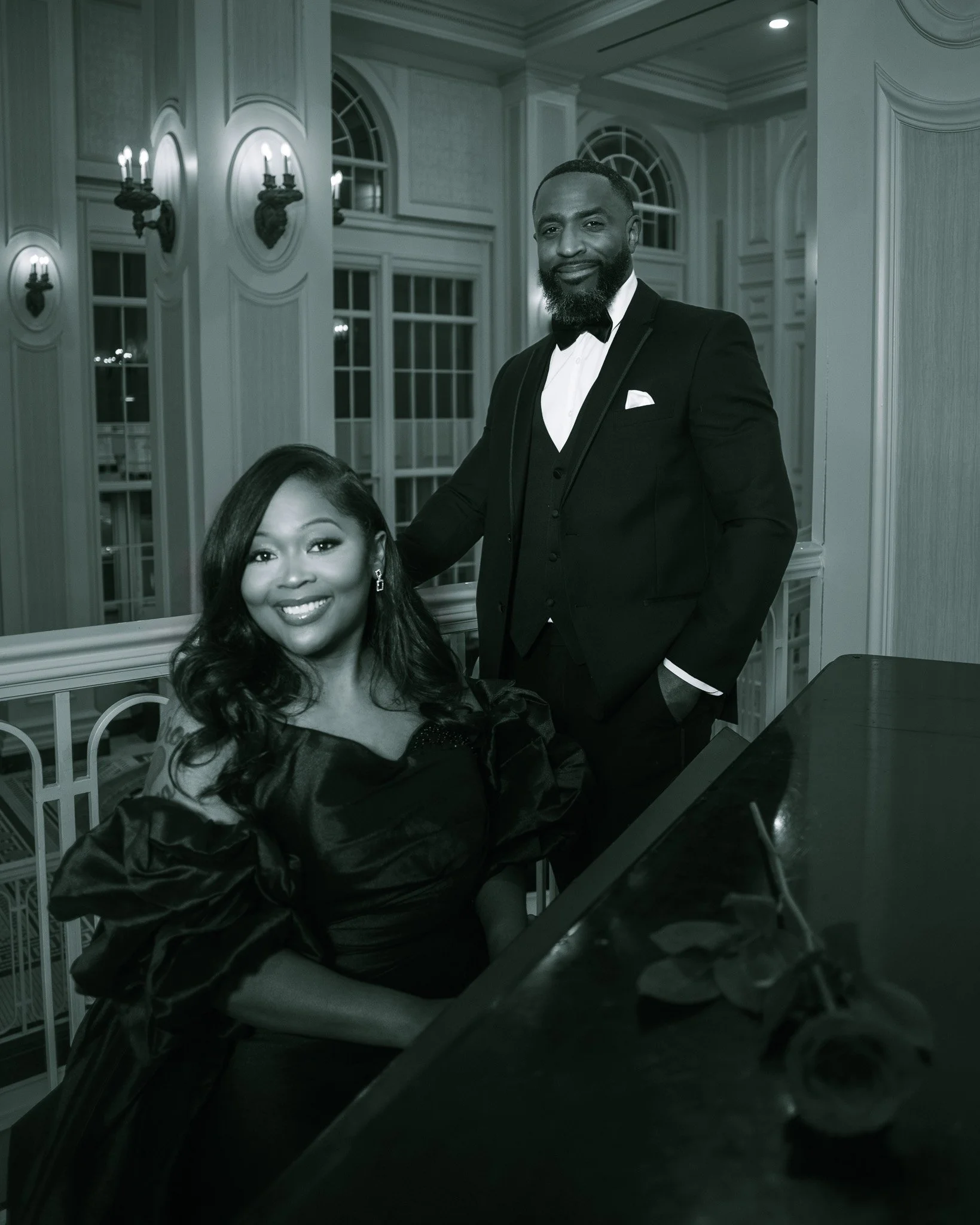 A smiling woman seated at a grand piano with a woman standing behind her and placing her hand on her shoulder, set in an elegant interior with large windows and ornate wall sconces.