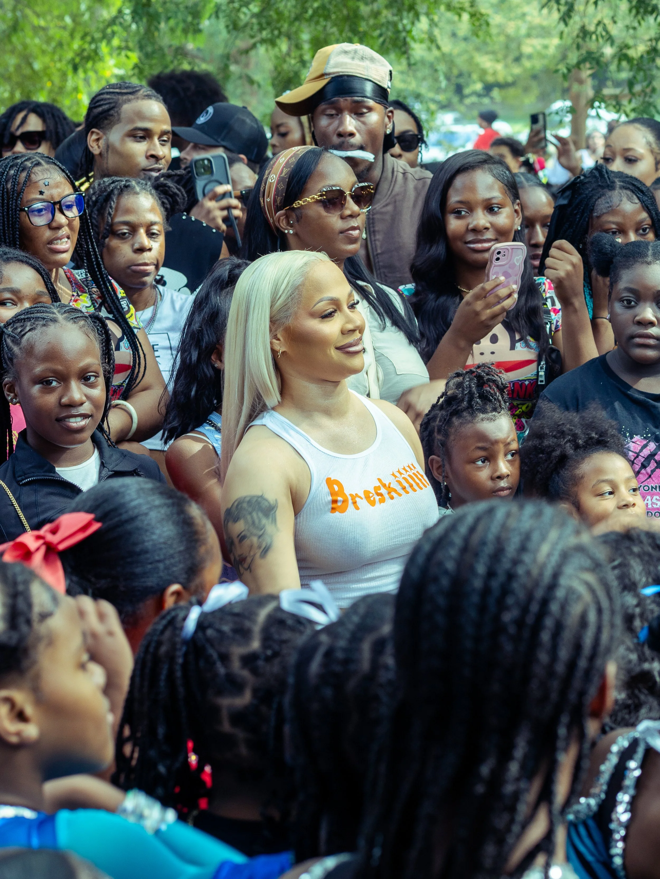 Crowd of diverse people, including children and adults, gathered outdoors, some taking photos or videos, with many smiling and looking in the same direction.