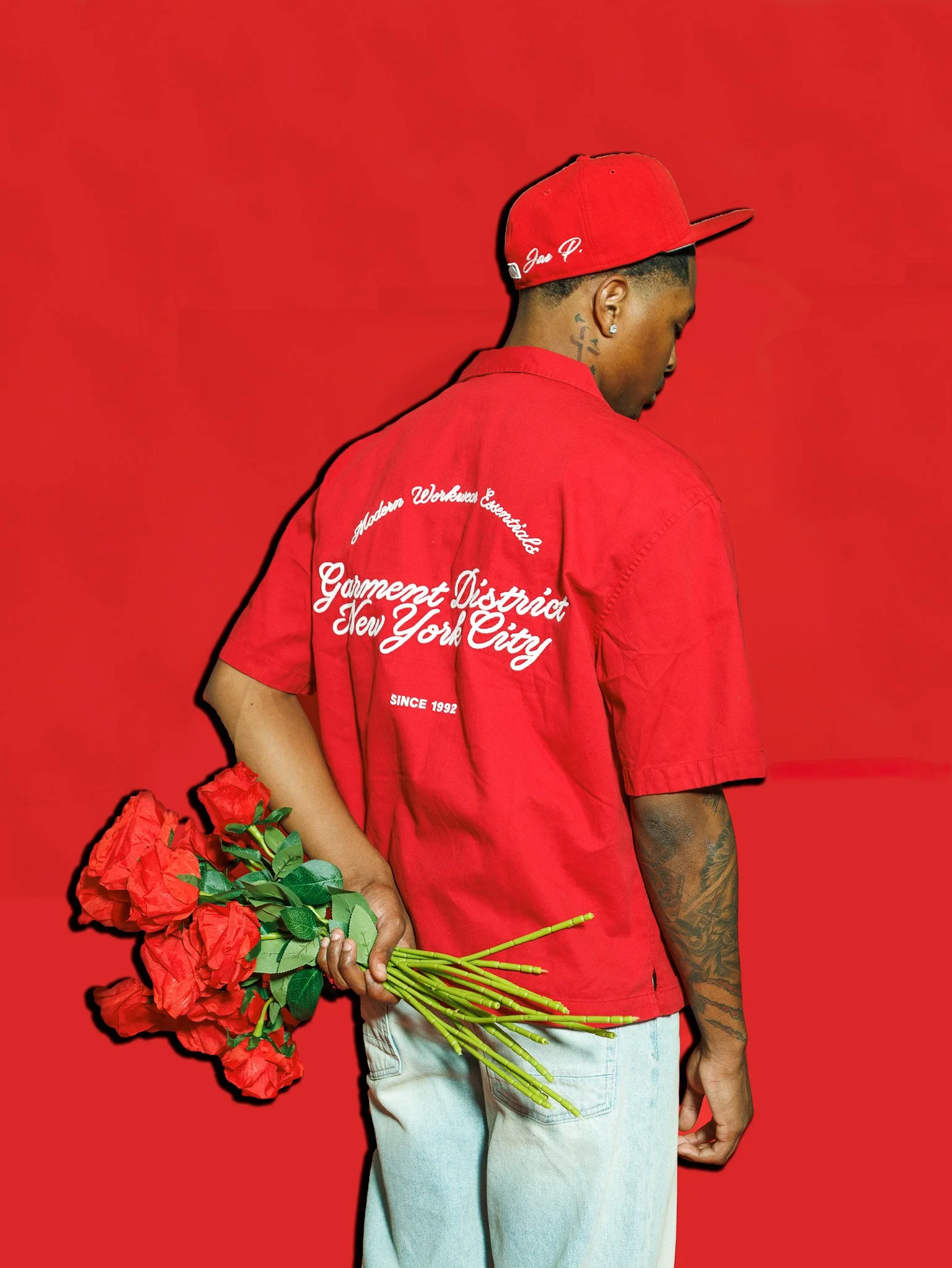 A man facing sideways holding a bouquet of red roses behind his back, dressed in a red shirt and red snapback hat, against a red background.