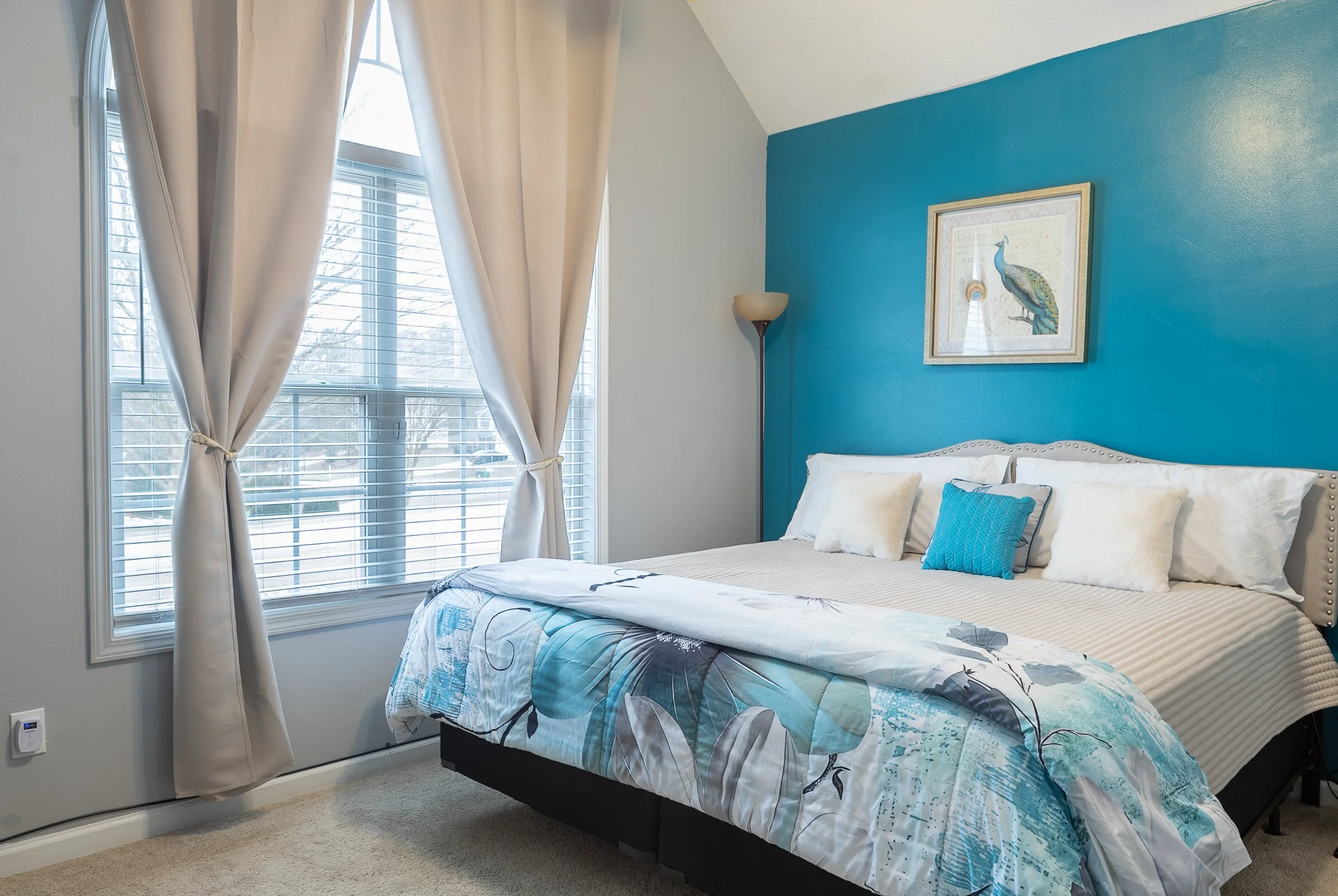 A bedroom with a blue accent wall, a bed with white and blue pillows, floral bedding, a framed peacock picture on the wall, a tall floor lamp, and a large window with cream curtains tied back, allowing natural light into the room.