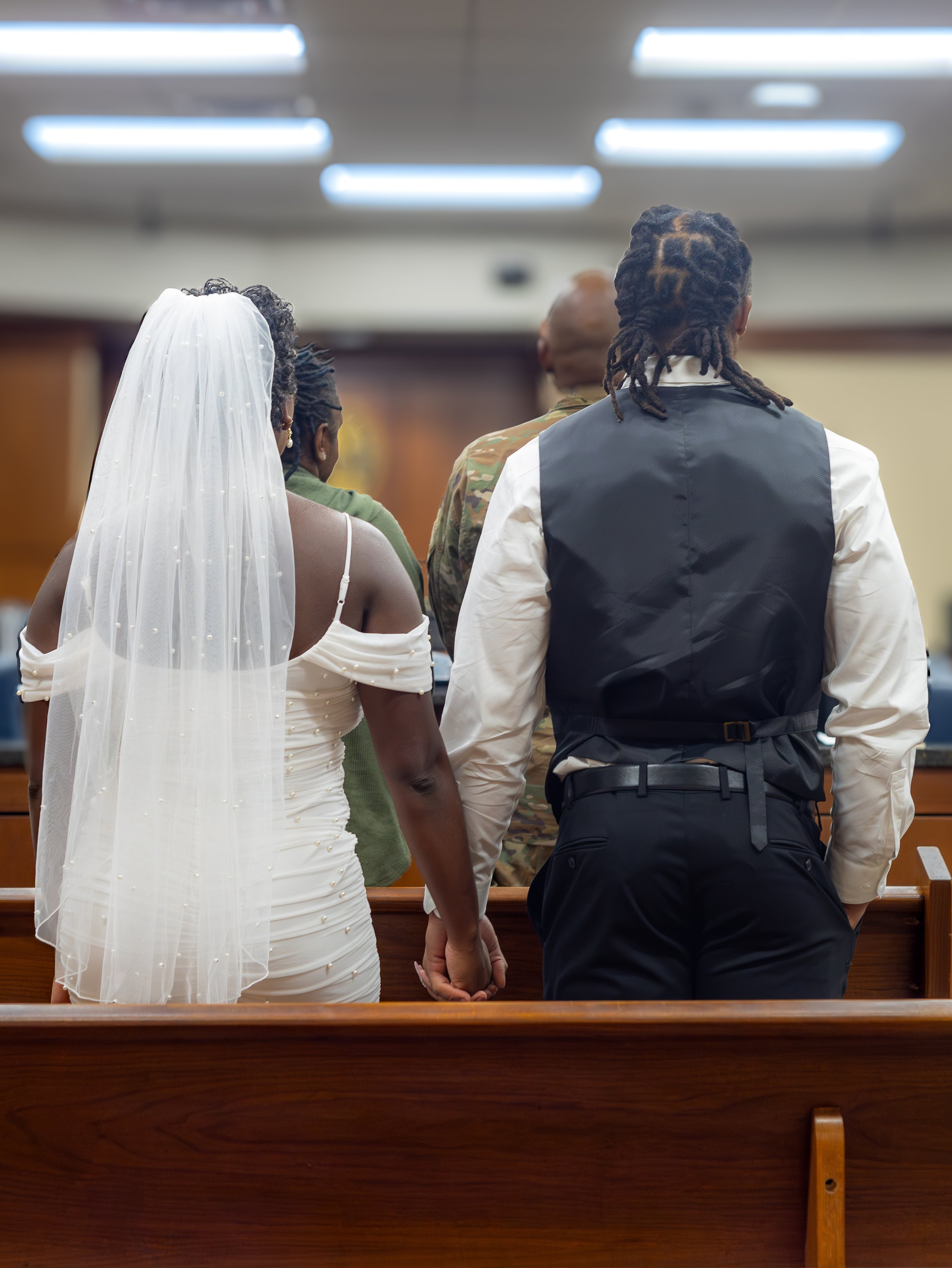 A bride and groom holding hands during a wedding ceremony in a church, facing forward, with witnesses and a judge present.
