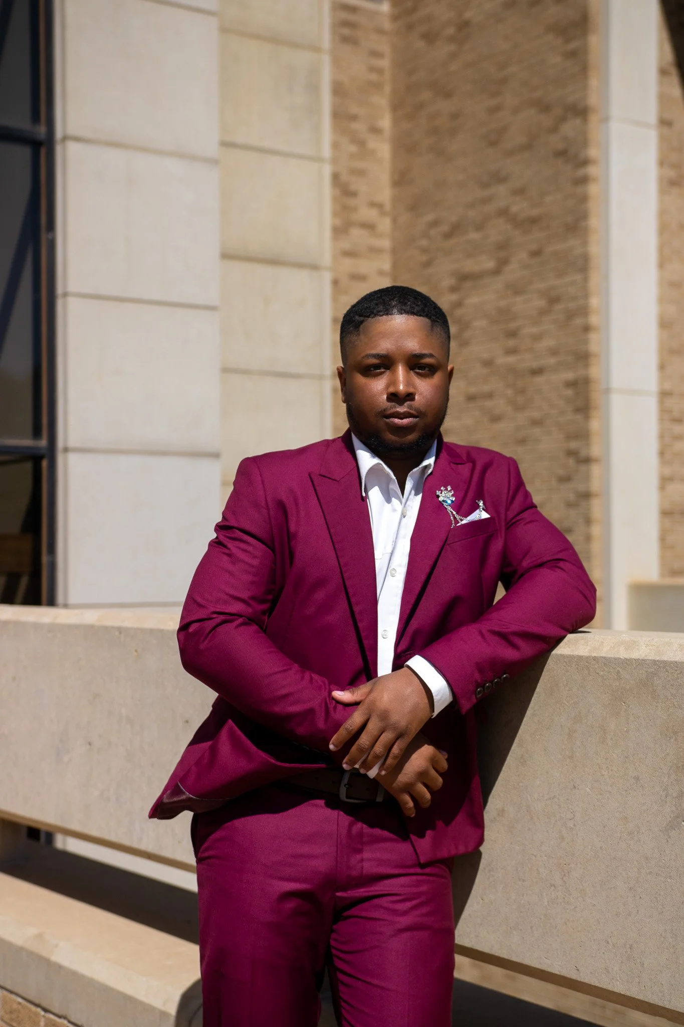 A man in a maroon suit and white shirt leaning against a concrete barrier outside a building with beige brick walls.