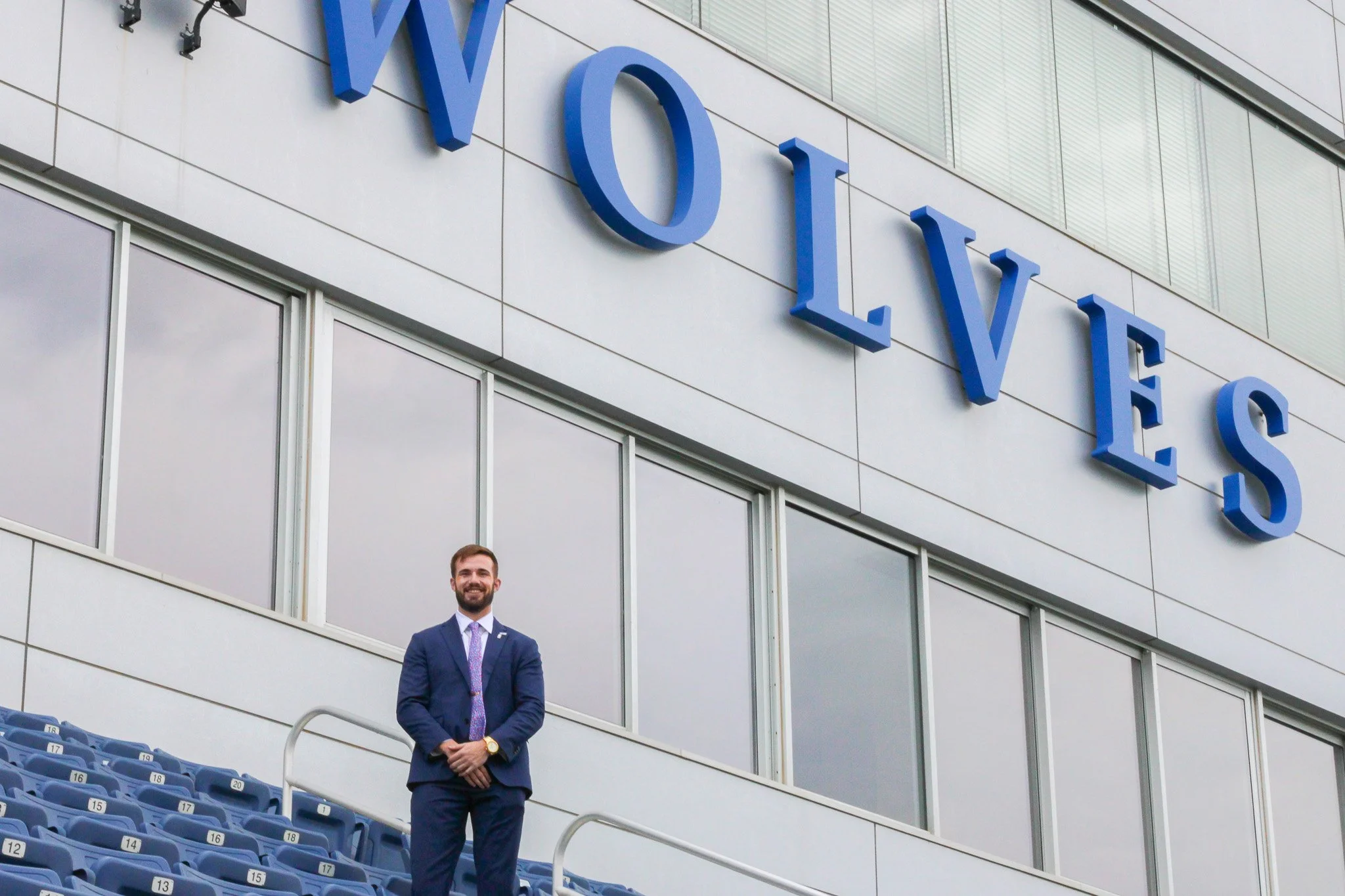 A man in a suit with a tie and a watch standing in front of a large building with a sign that reads 'WOLVES' in big blue letters.